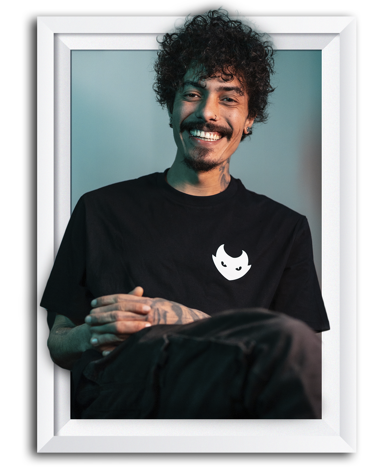 Smiling person with curly hair wearing a black shirt featuring a white cartoon logo inside a white frame.