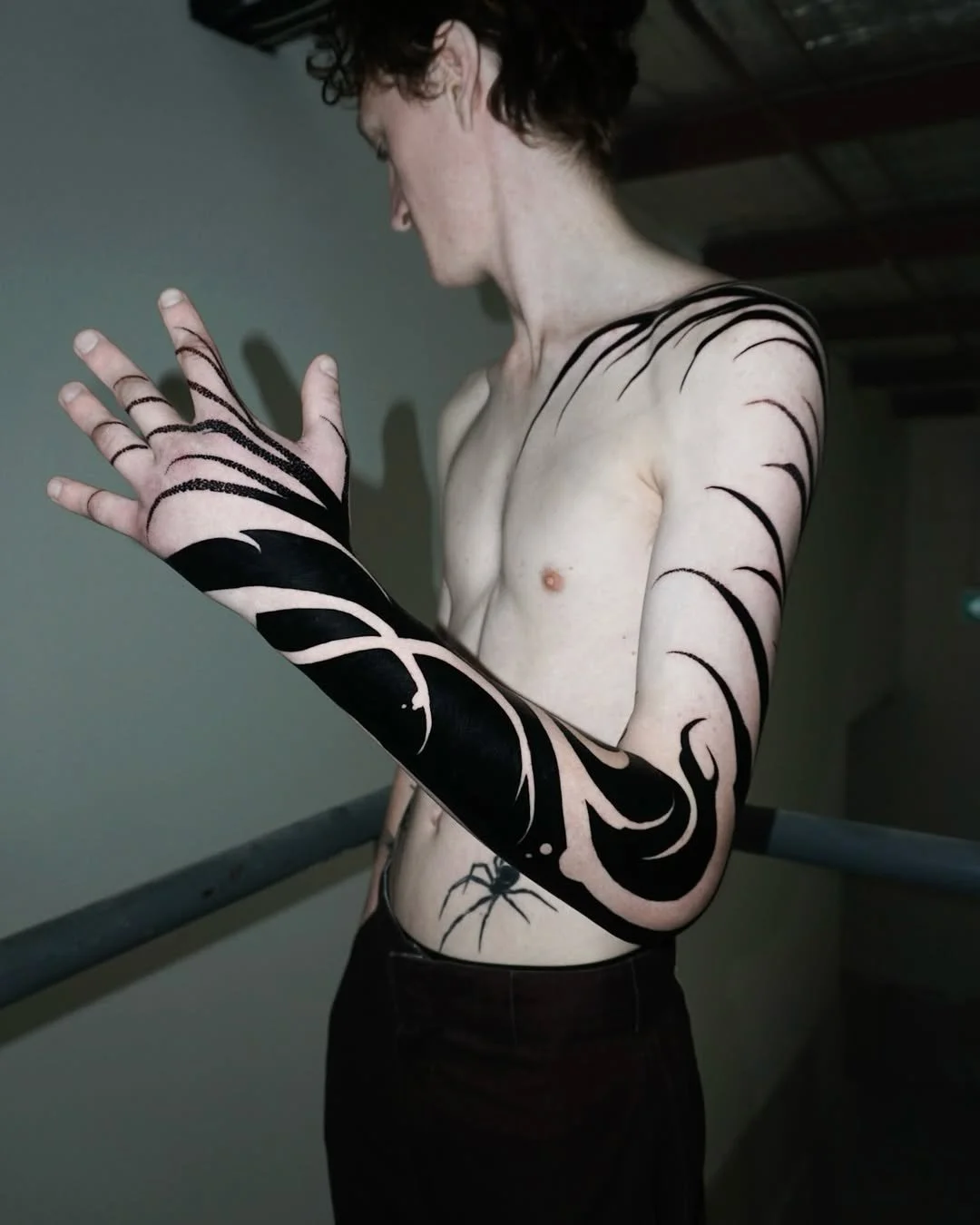 A topless person with short curly hair and extensive black tattoo artwork covering their arms and side, standing indoors near a railing, looking at their hand.