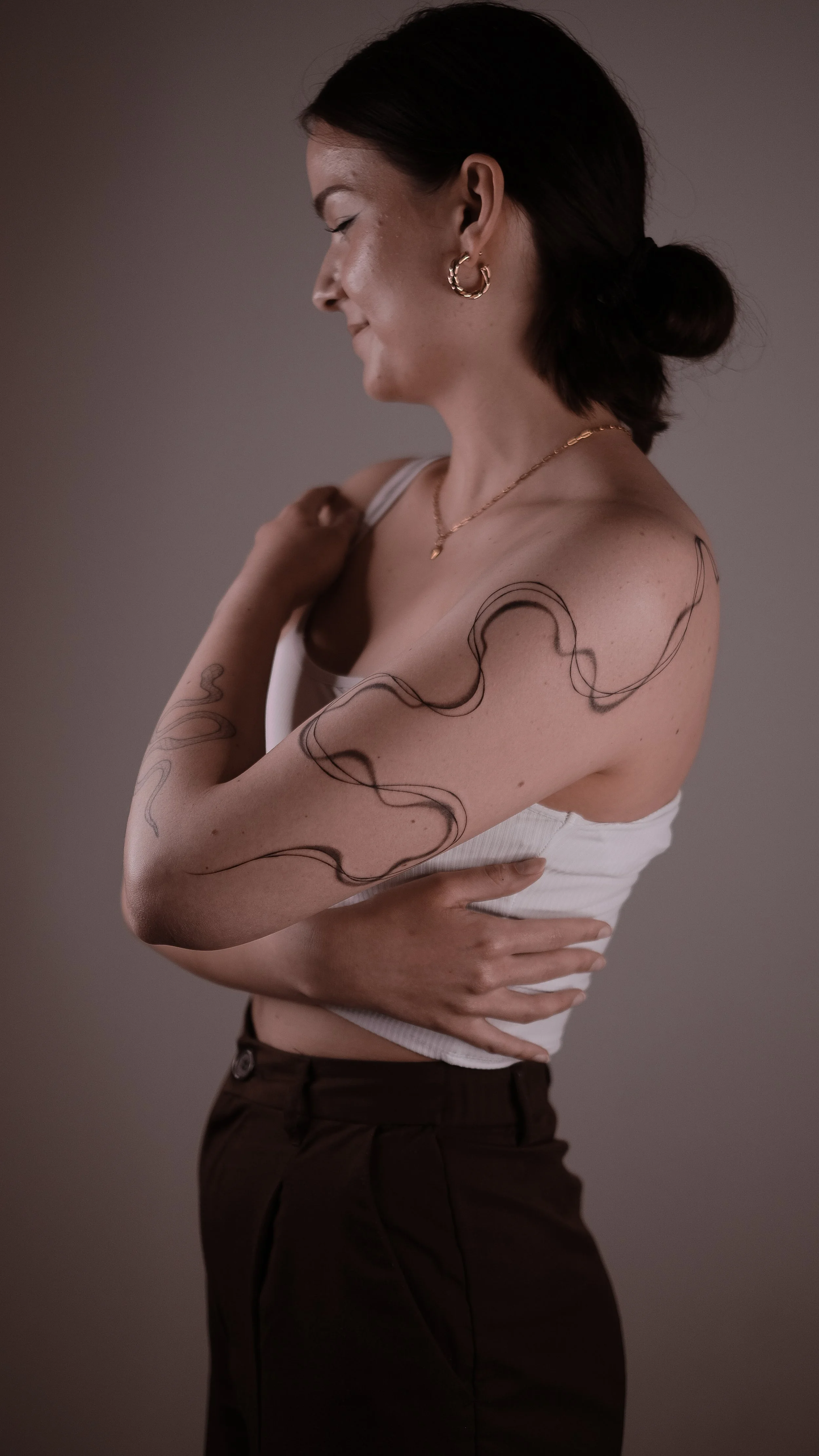 Woman with black hair in a bun, wearing hoop earrings and a white top, displaying a snake-like tattoo on her arm.