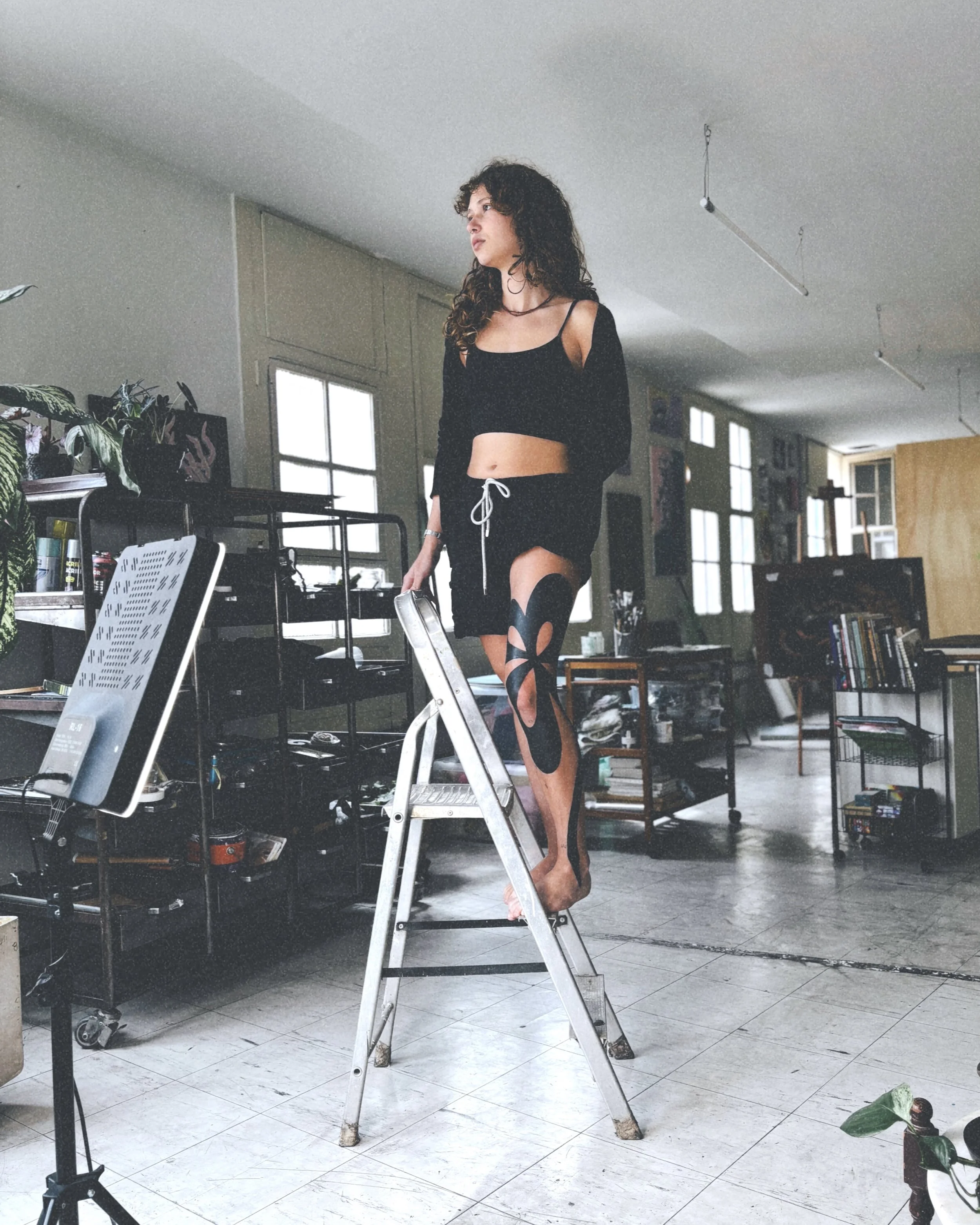 A woman with curly hair standing on a ladder in an art studio, wearing a black crop top, shorts, and eye-catching cut-out tights, surrounded by art supplies and books.