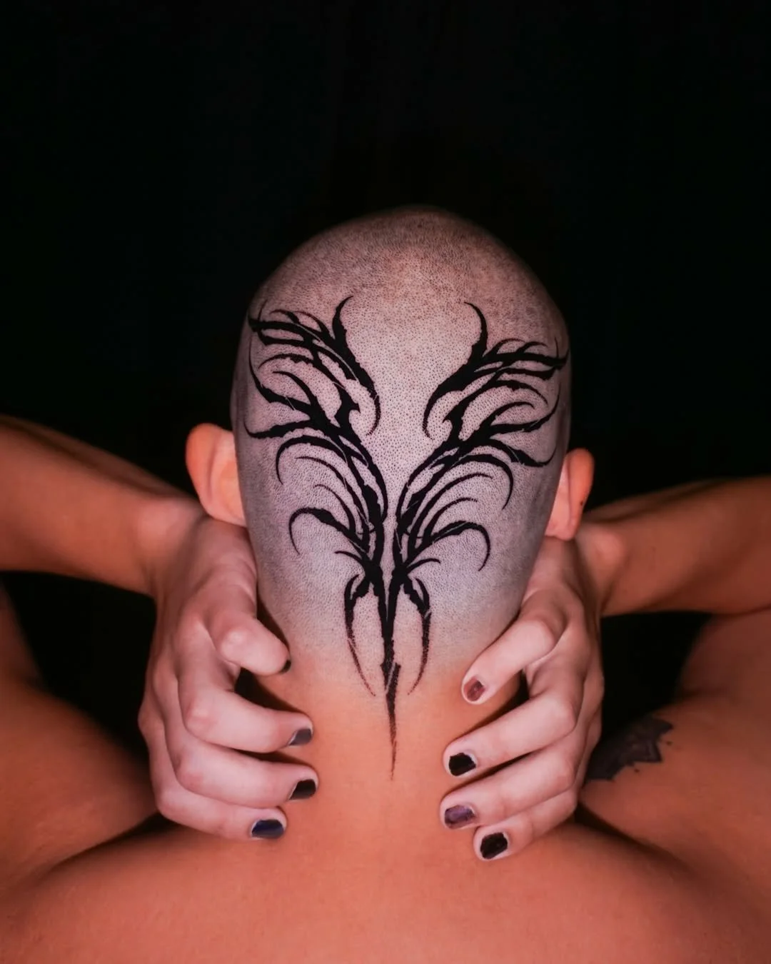 A person with a bald head featuring a large, black tribal tattoo design in the center of their scalp, with hands holding their head. The background is black.