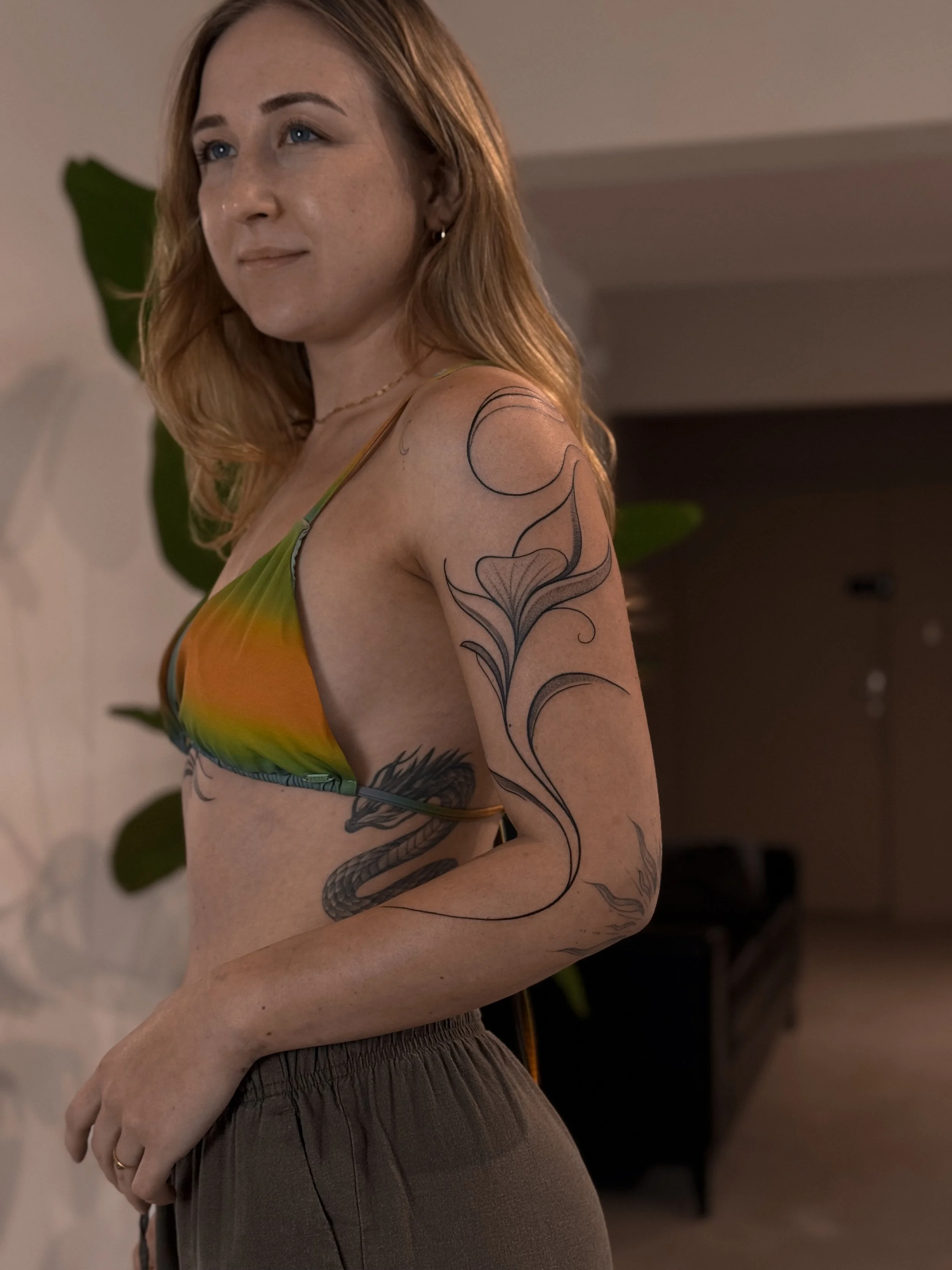 A woman with red hair and tattoos on her arm and side, wearing a colorful bikini top and brown pants, standing indoors with plants and furniture in the background.