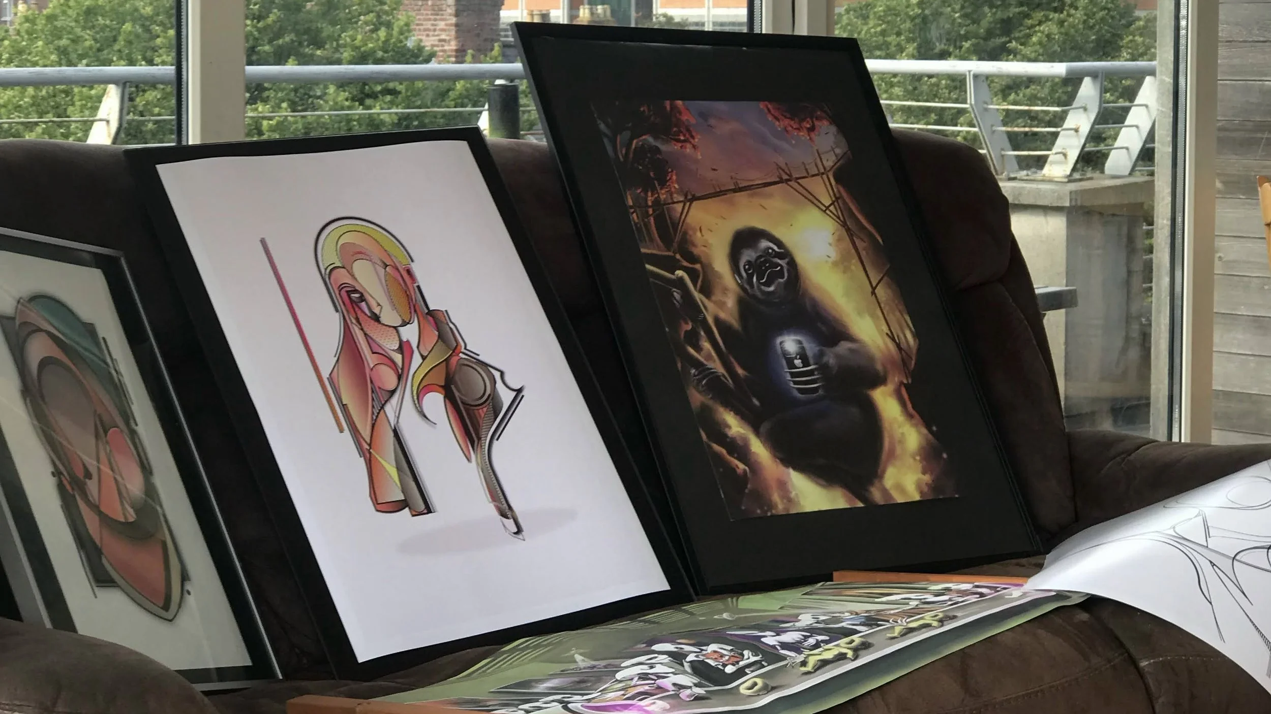 Framed abstract and surreal artwork leaning against a couch, with bright window in the background.