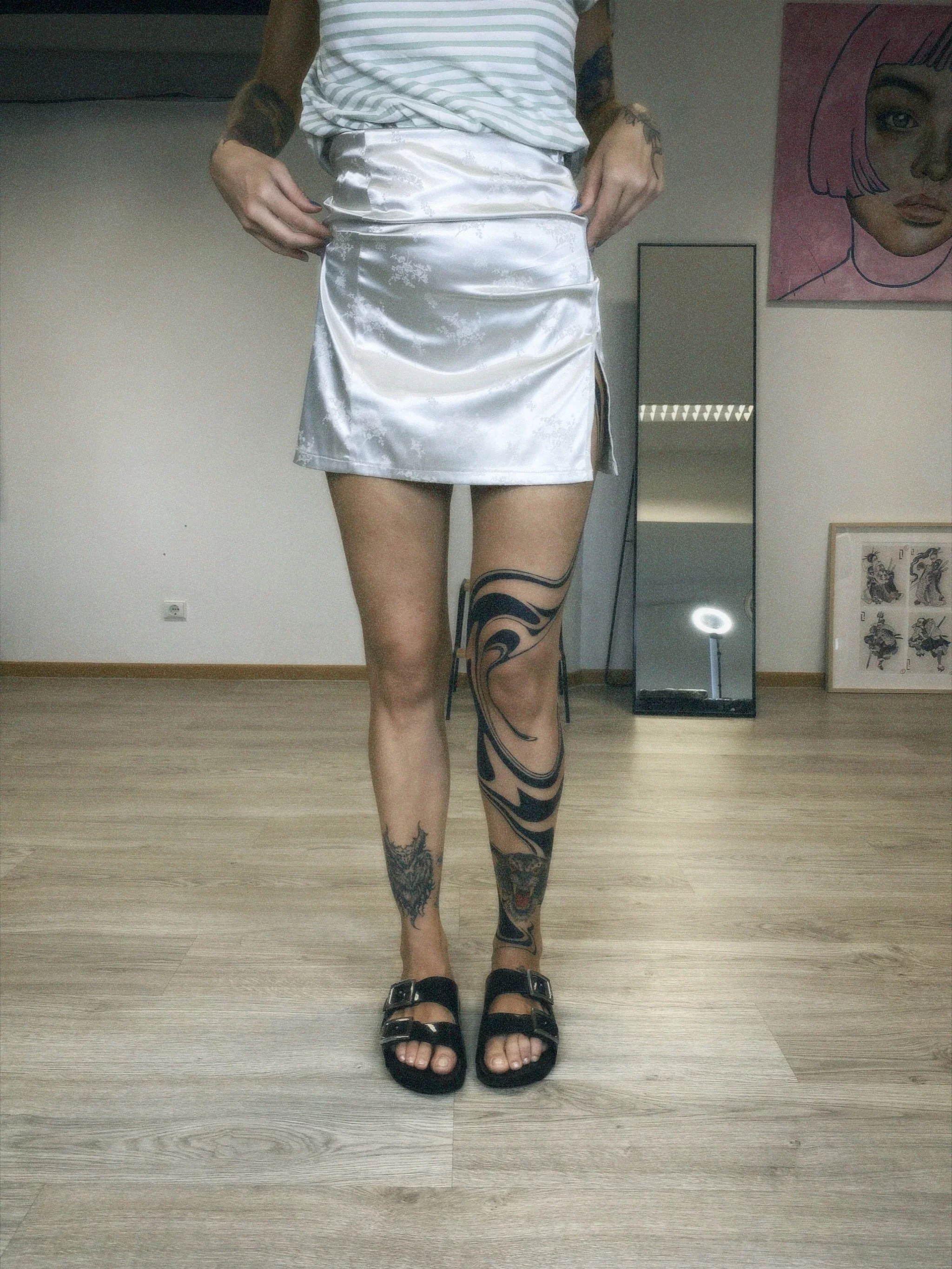 Person wearing a white satin skirt, black sandals, and has tattoos on legs, standing on a wooden floor in front of a mirror.