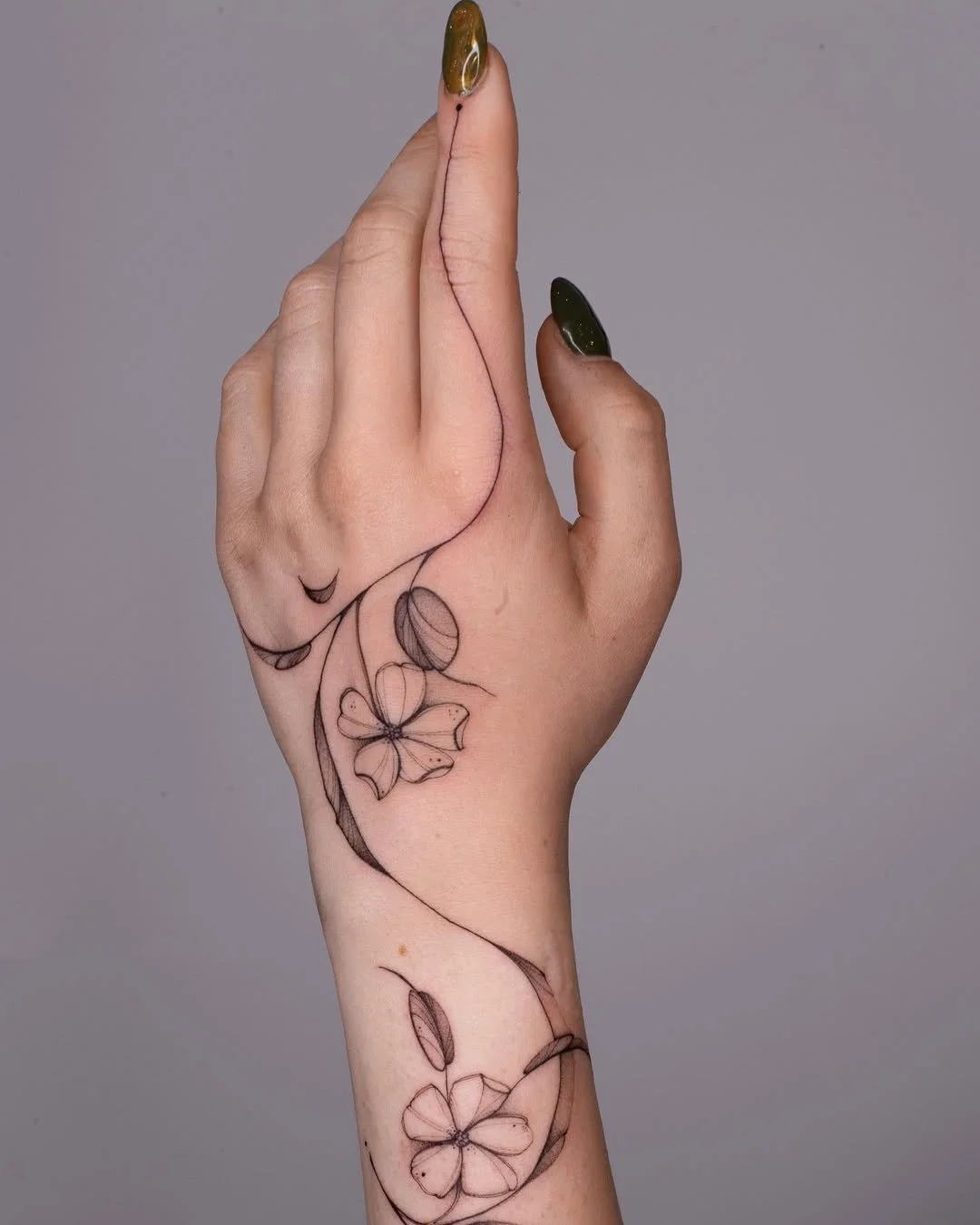 A person's hand with floral tattoos on the wrist and forearm, held in a prayer-like position with green and gold nail polish, against a plain gray background.