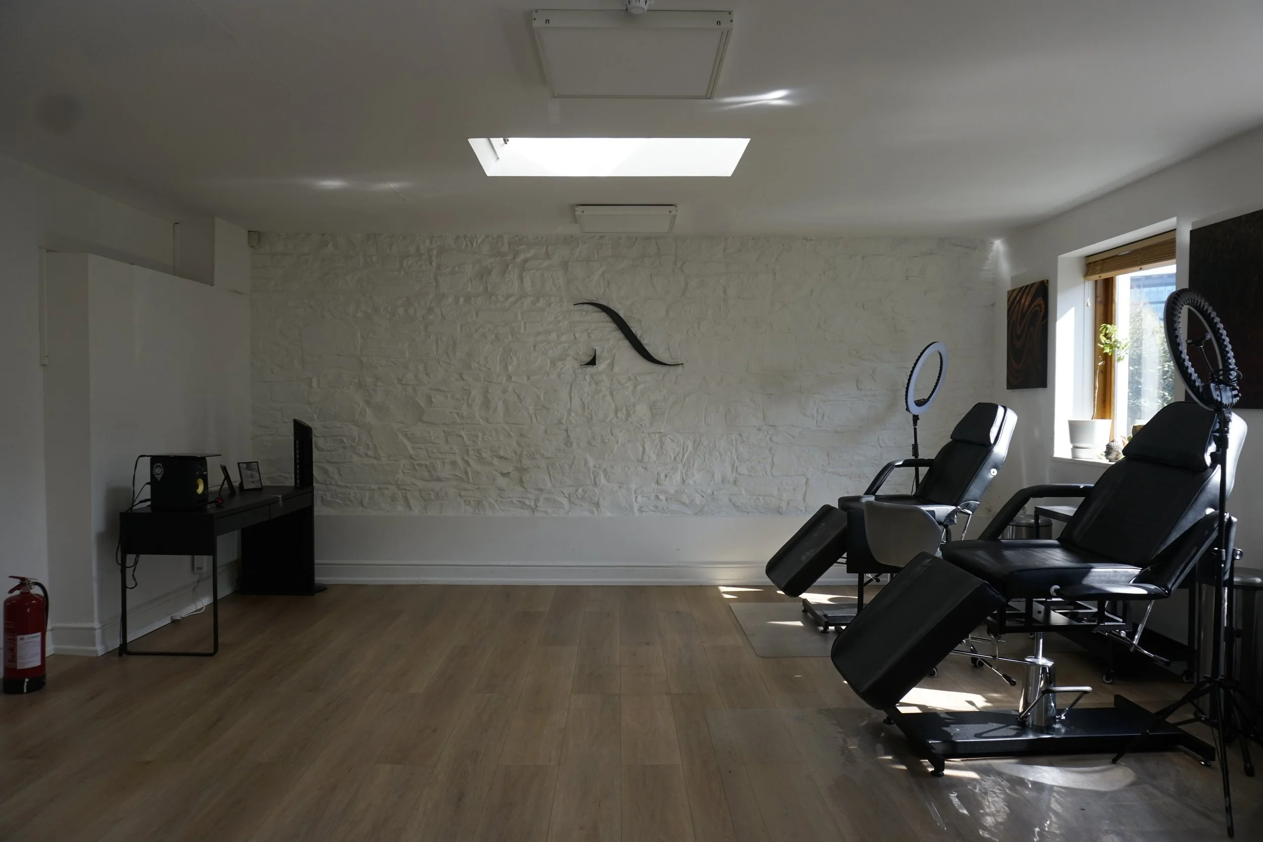 A tattoo studio with two black tattoo chairs, ring lights, a wooden floor, and a skylight. The room has a white brick wall, a small desk with equipment, and a fire extinguisher.