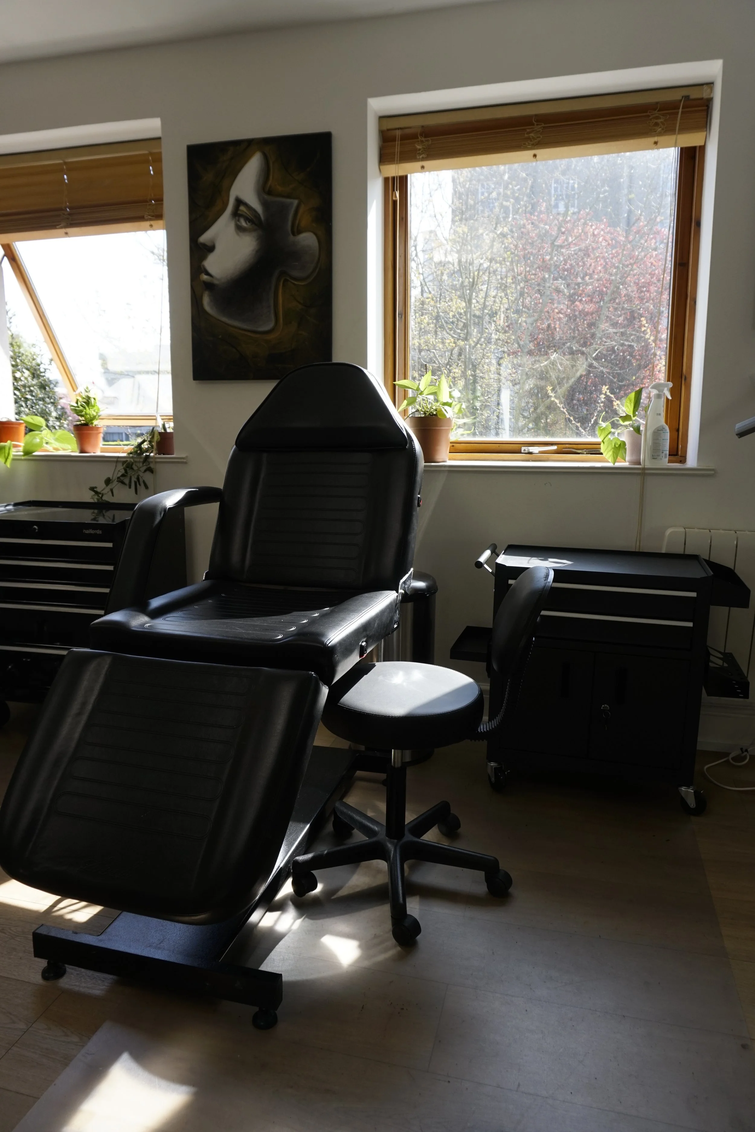 A tattoo studio setup with a black reclining chair on a wooden floor, adjacent to a black rolling stool. A wall features a portrait painting. Two windows with wooden blinds provide natural light, and houseplants sit on the window sill.