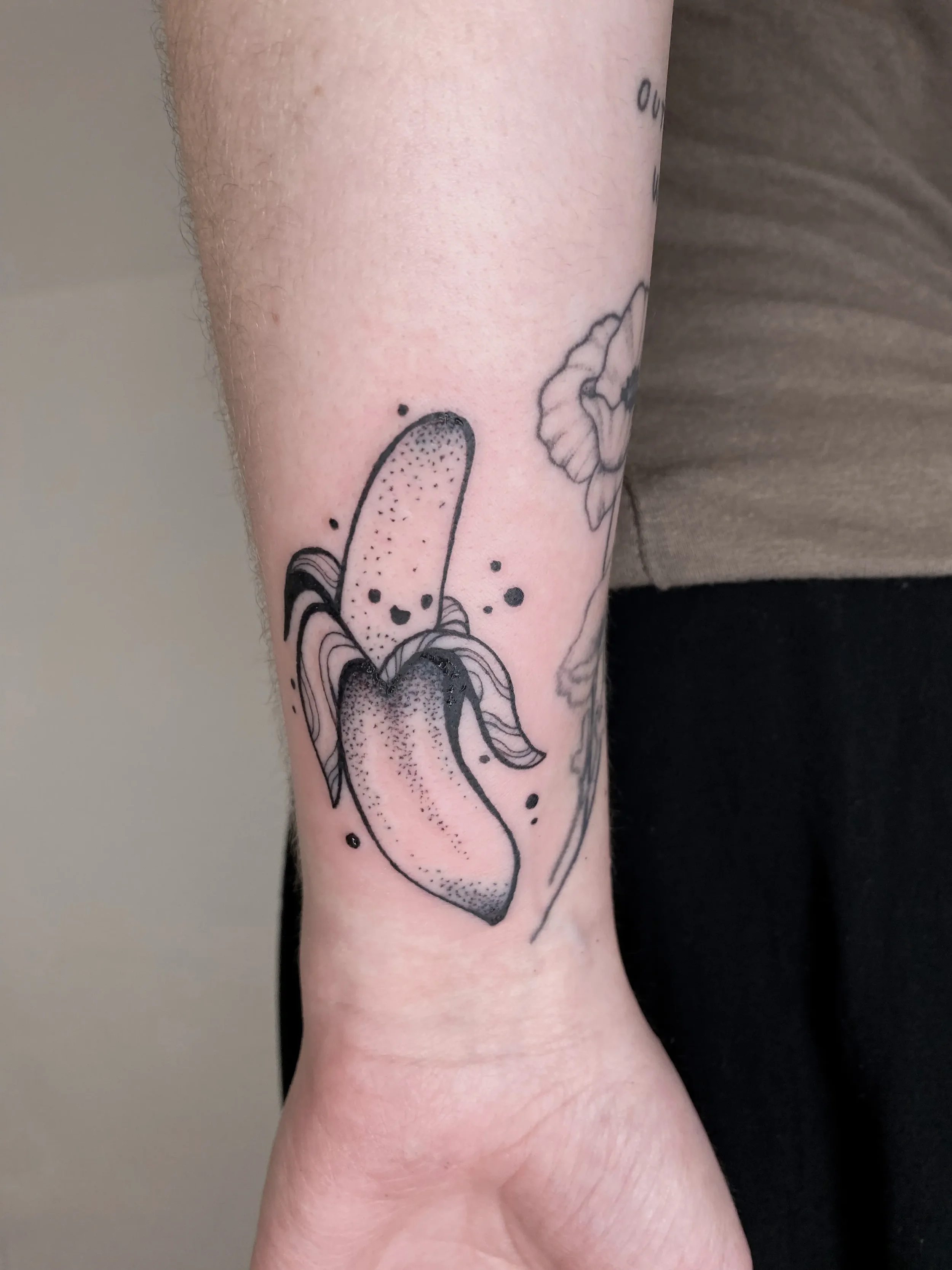 A black ink tattoo of a banana on a person's forearm, with stylized lines and dot shading, surrounded by small black ink splatters.