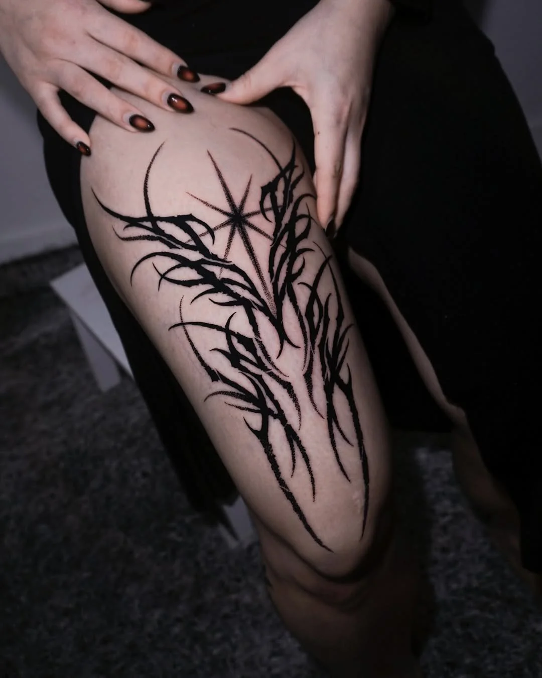 A person's thigh with a black tribal-style tattoo of a tree or branches and a central star. The person has manicured nails painted with a dark, shiny color.
