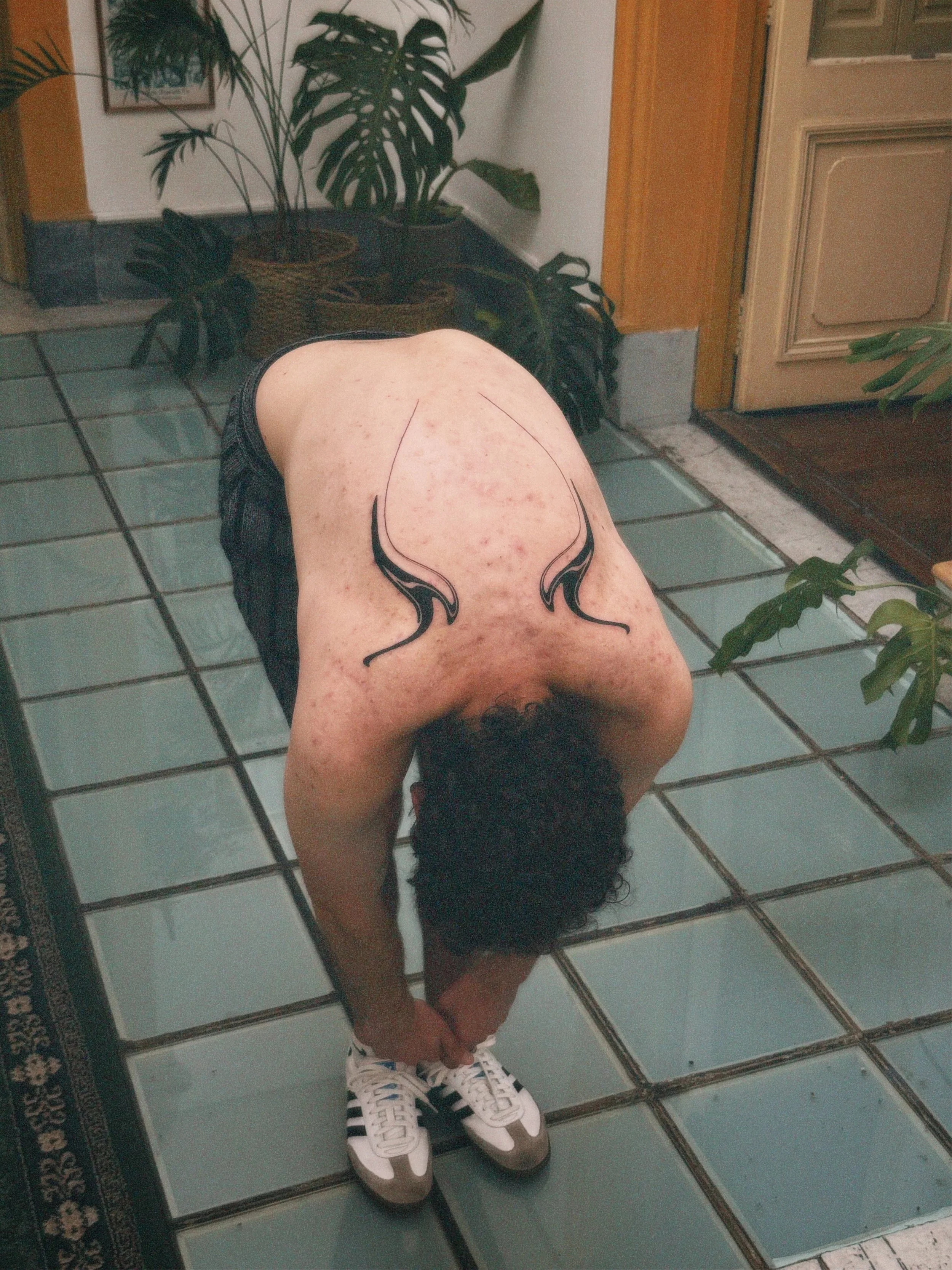 Person with tattoo on back bending down to tie shoelaces, in an indoor setting with plants and tiled floor.