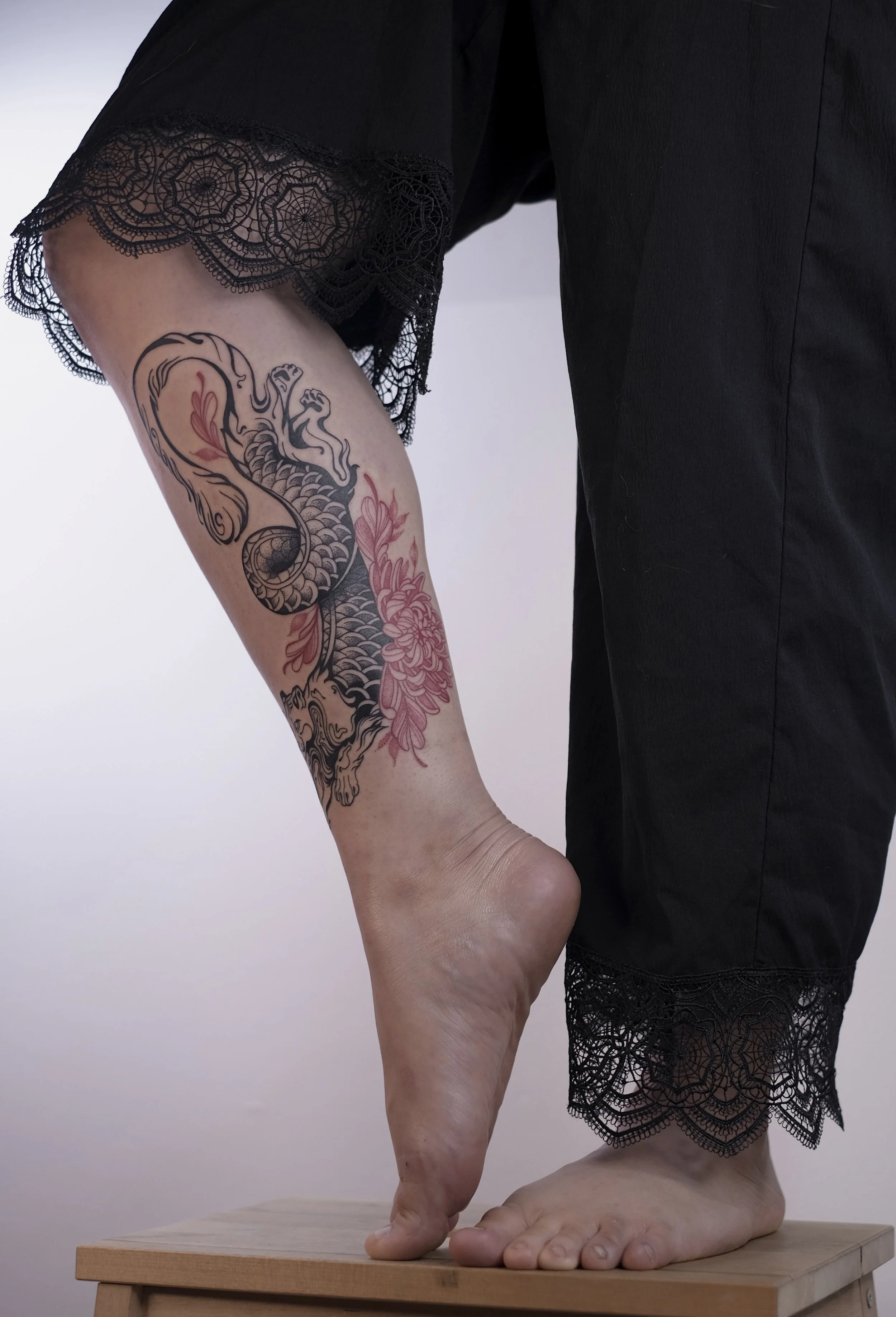 Close-up of a person's leg with a colorful dragon tattoo, wearing black lace-trimmed pants, standing on a wooden platform.