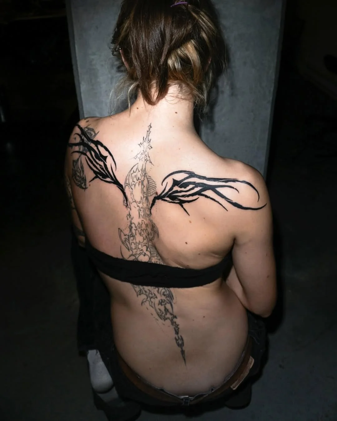 A woman with shoulder-length hair, seen from behind, has a large tattoo on her back featuring a dragon with wings, with the tattoo extending down her spine.
