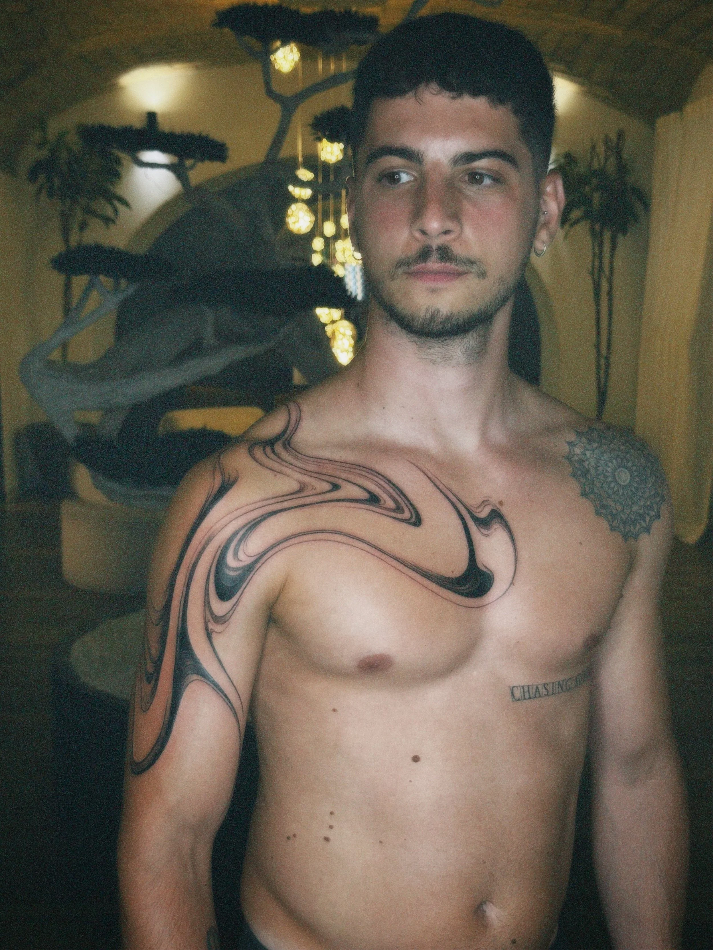 A young man with short dark hair and facial hair, shirtless, showing tattoos on his shoulder, chest, and upper arm, standing indoors with warm lighting and decorative plant art in the background.