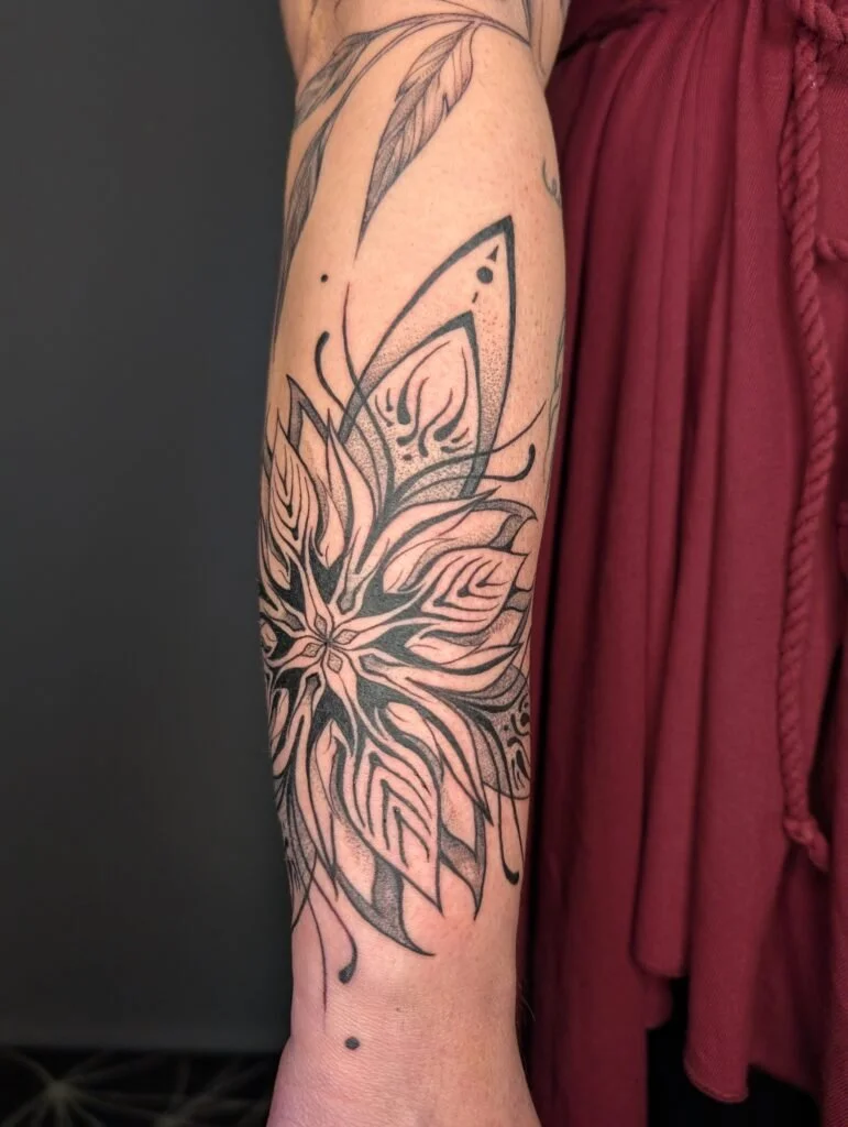 Black and gray floral tattoo on person's forearm, featuring a large flower with detailed petals and leaves.