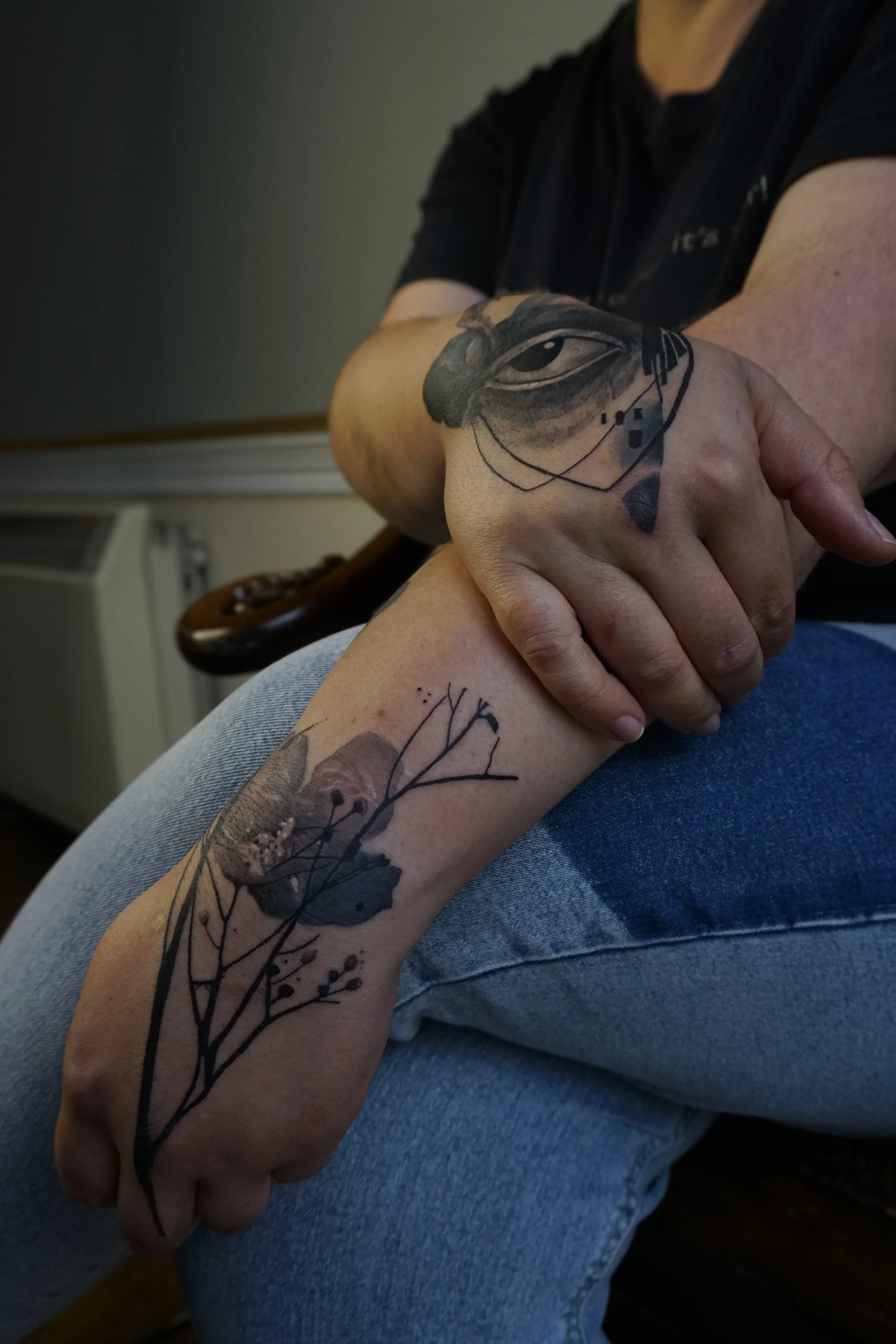 A person with a tattoo of an eye and face on their right hand and a floral design on their left wrist. The person is sitting with arms crossed, wearing a black shirt and blue jeans.