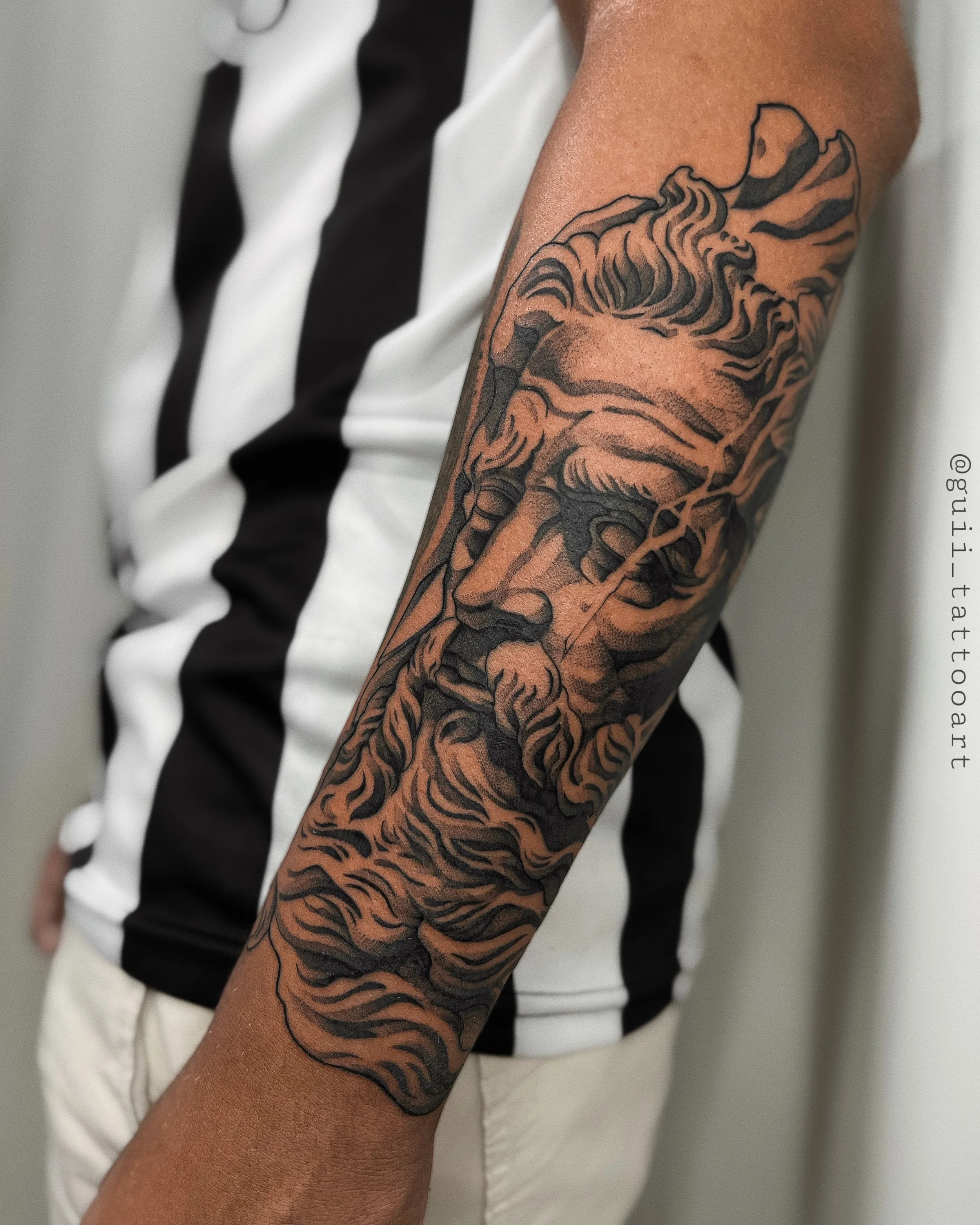 Forearm tattoo of a classical Greek-style face with flowing hair and beard.