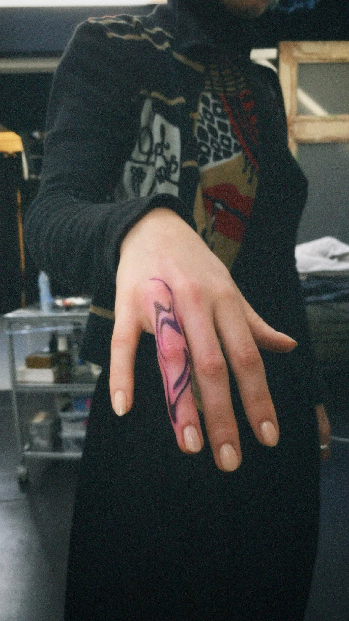 A person shows a colorful, abstract tattoo on their left middle finger. They are wearing a black long-sleeve shirt with a graphic design. The background appears to be a room or studio with various objects and furniture.