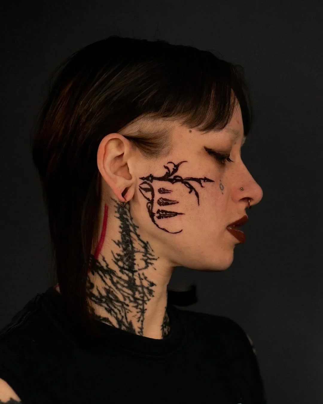 Side profile of a person with facial and neck tattoos, black layered hairstyle, and facial piercings, against a dark background.