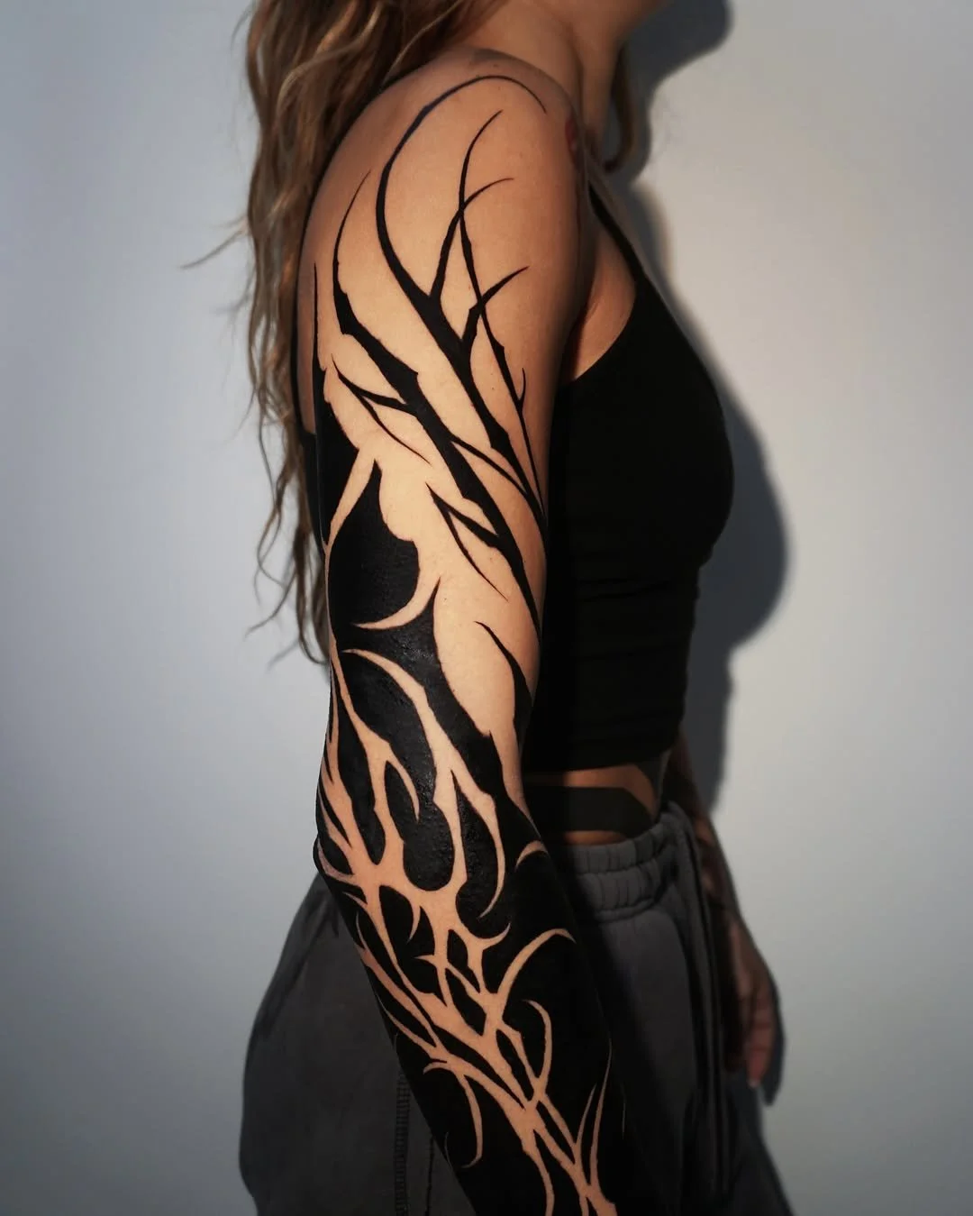 A woman with a tribal-style black tattoo covering her arm and shoulder, wearing a black sleeveless top and gray sweatpants.