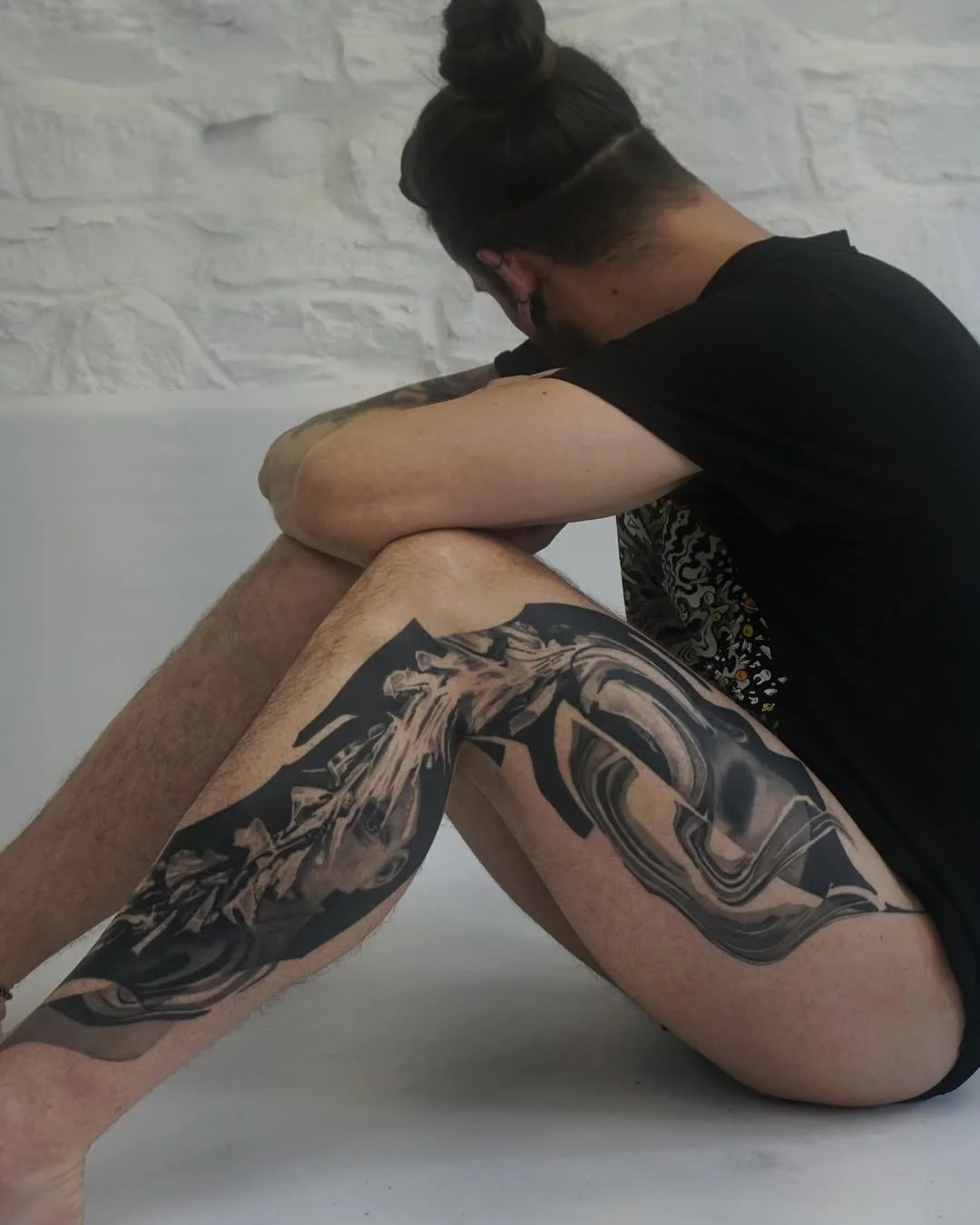 Person with dark hair in a bun, wearing a black shirt, sitting on the floor with their head resting on their knees, tattoos visible on their arms and legs, in front of a white brick wall.
