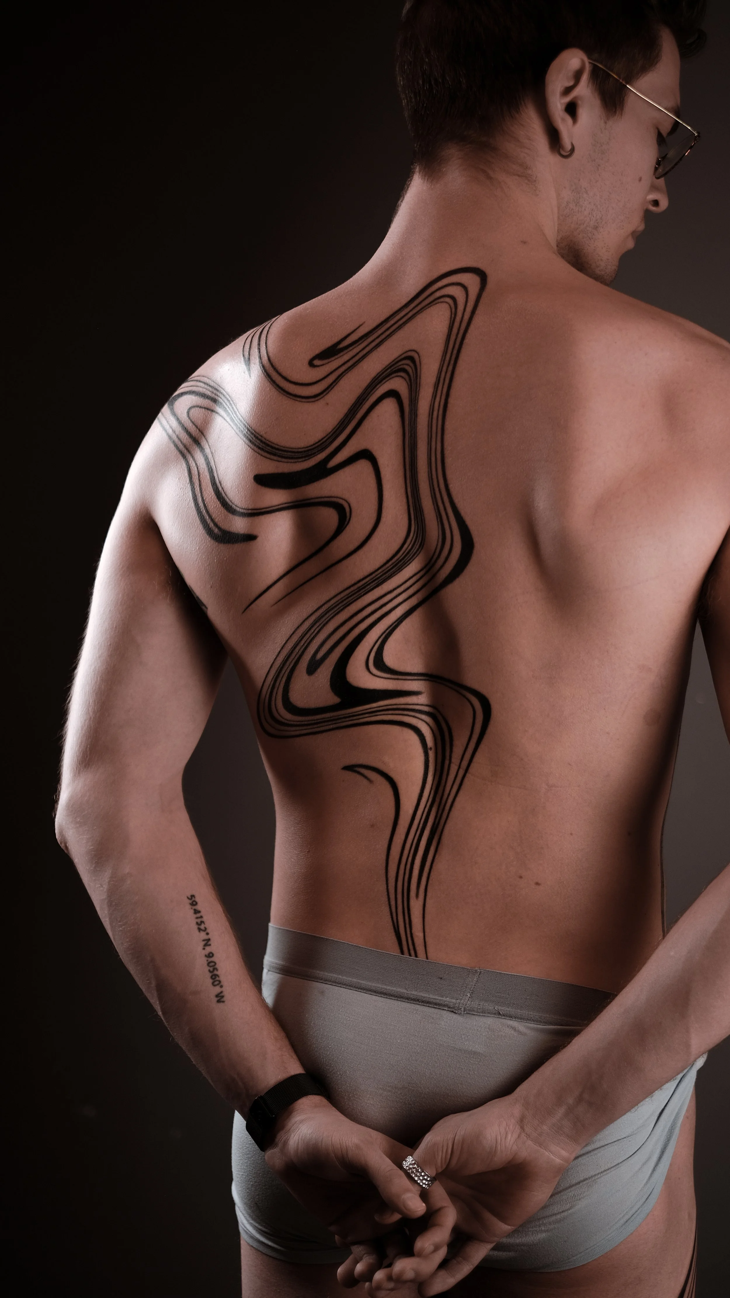 Man with abstract black tattoo on his back, wearing glasses and gray underwear, facing away from the camera with hands behind his back.