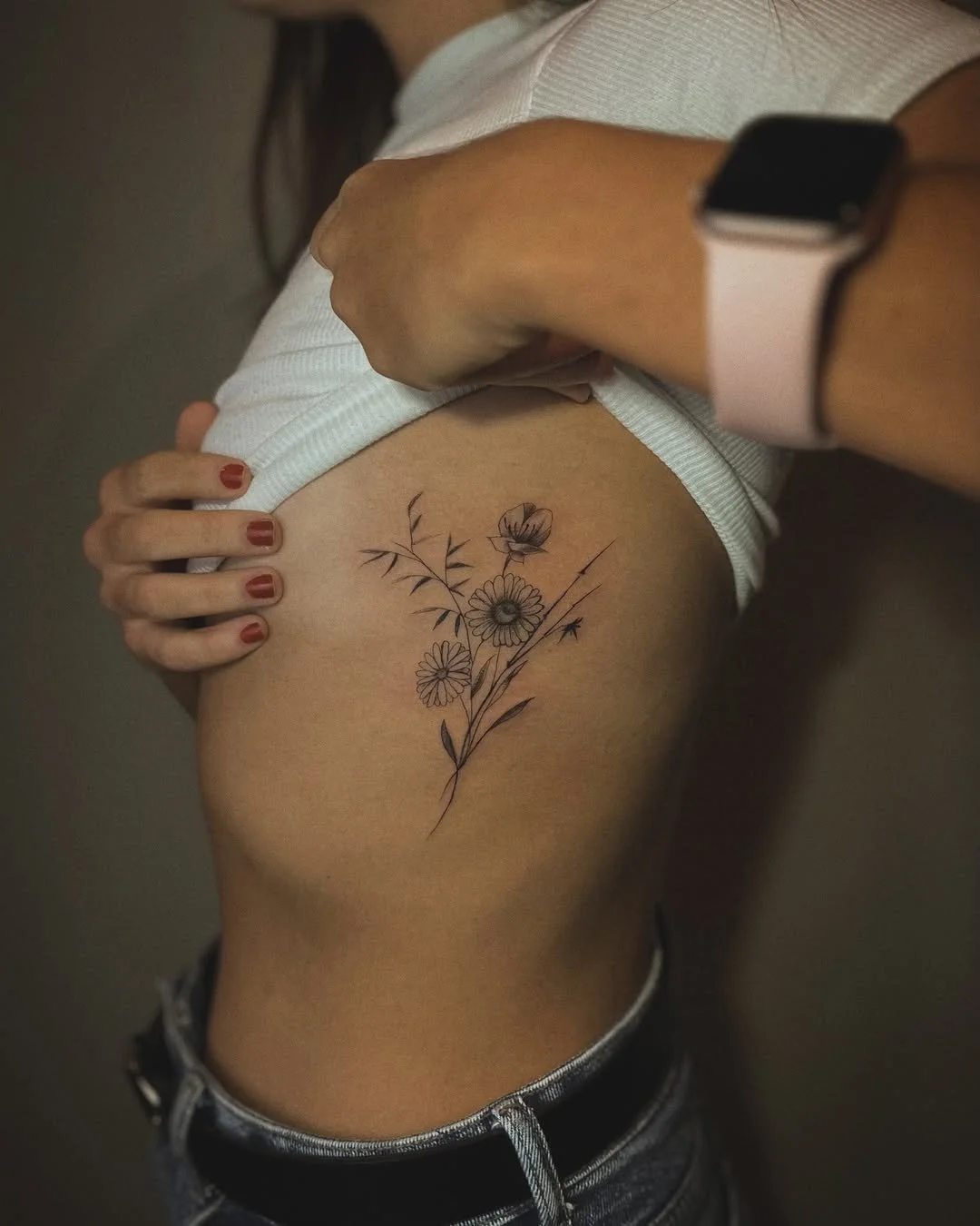 A woman is lifting her white shirt to reveal a floral tattoo on her left side, near her ribs, with her left hand resting on her waist and her right hand gently lifting the shirt. She has red nail polish and a smartwatch with a pink band on her right 