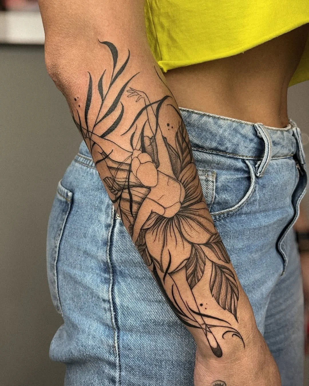 Close-up of a person's arm with a large floral and abstract tattoo, wearing light blue jeans and a yellow top.