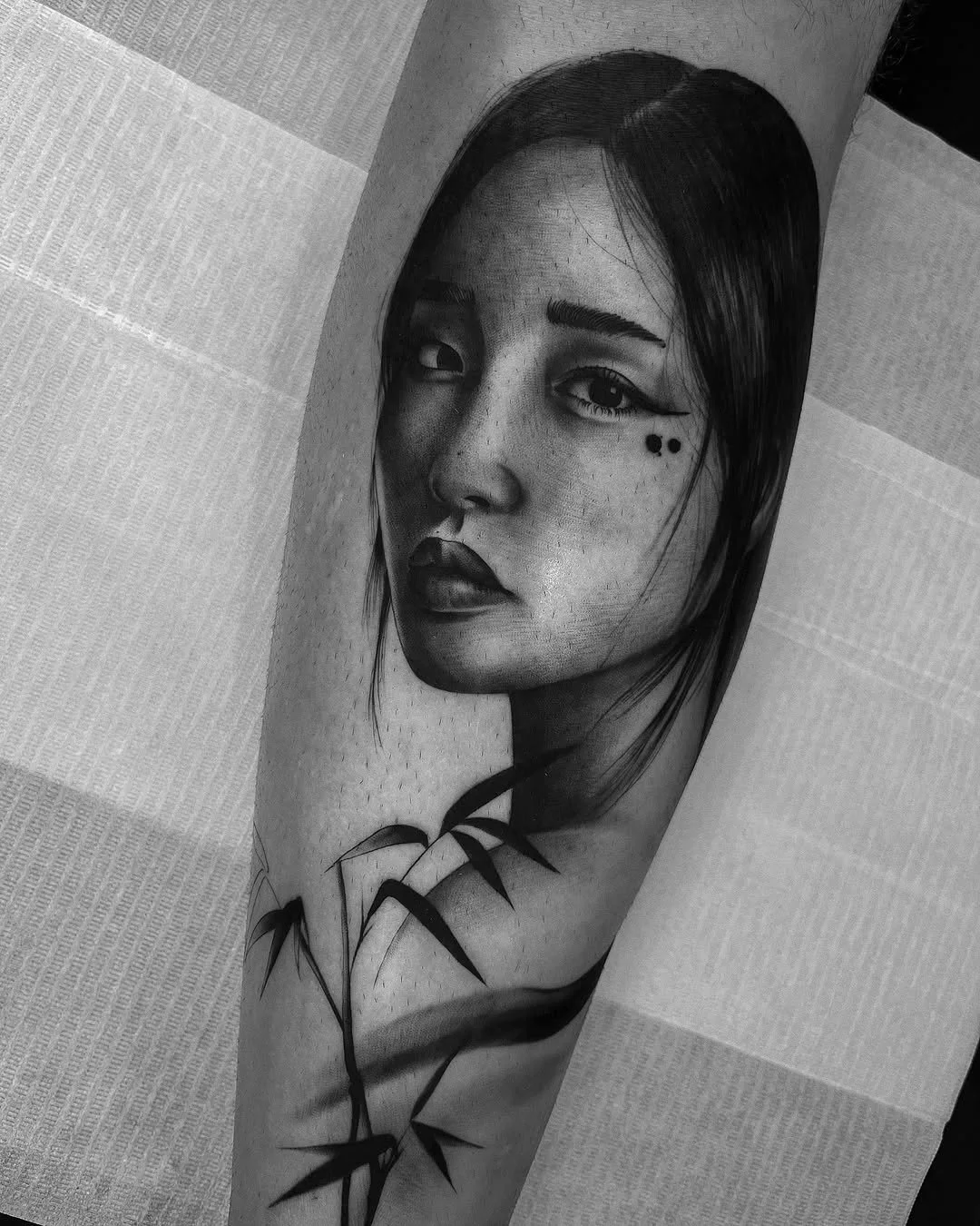 Black and white tattoo of a woman's face with long hair and bamboo leaves on skin.