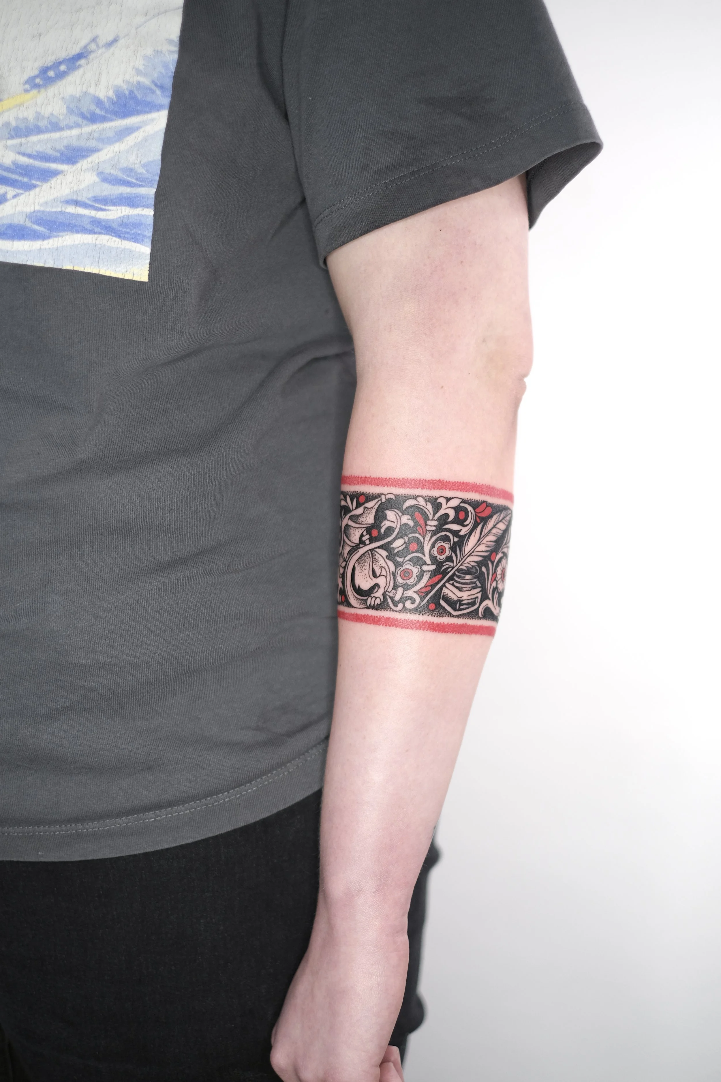 Person wearing a black T-shirt and black pants with a detailed black tattoo on their left forearm featuring floral and skull designs, with red borders around the tattoo.