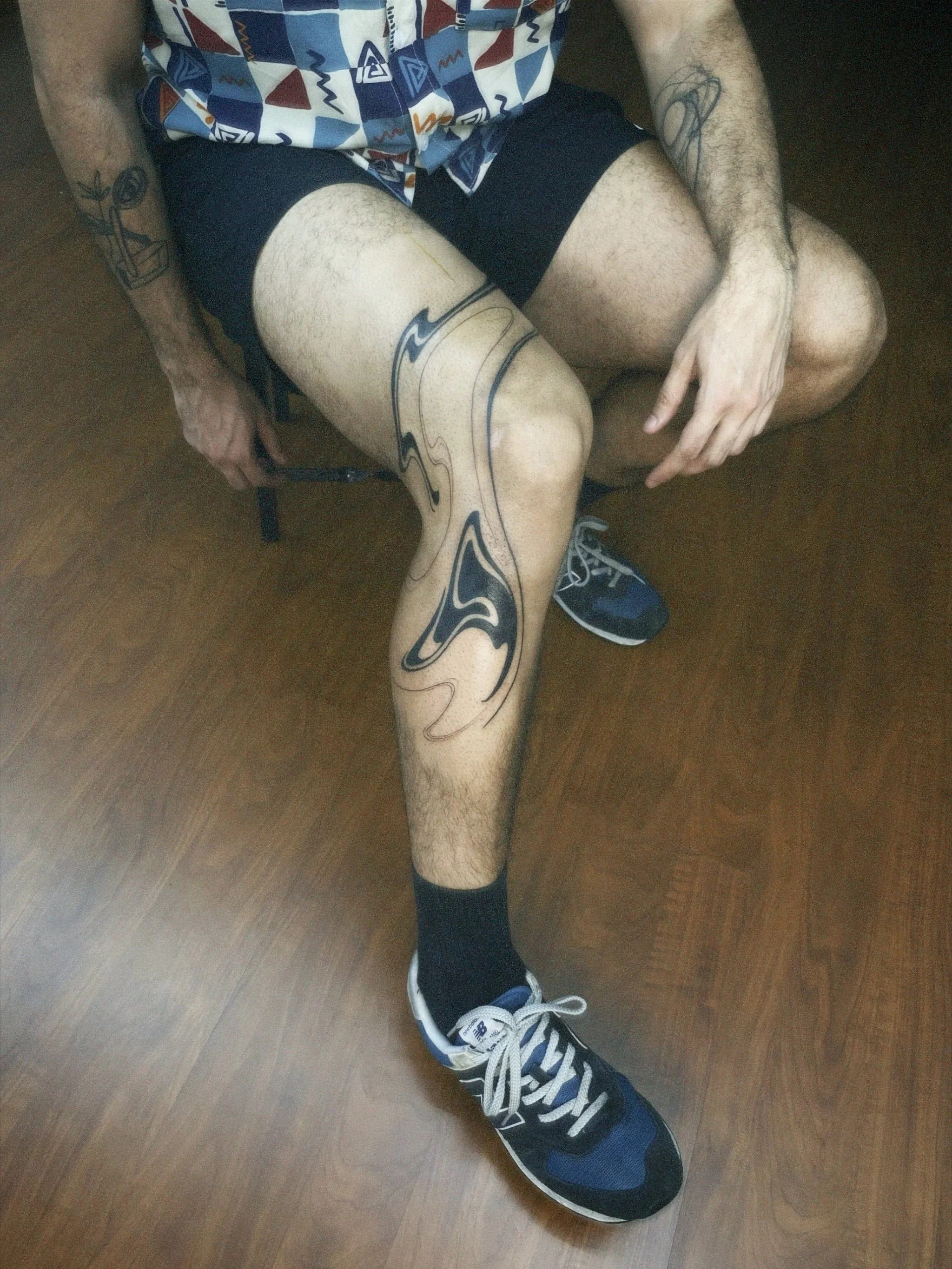 Person sitting on a chair with a patterned shirt, black shorts, and artistic tattoos on their legs and arms, wearing blue and white athletic shoes.