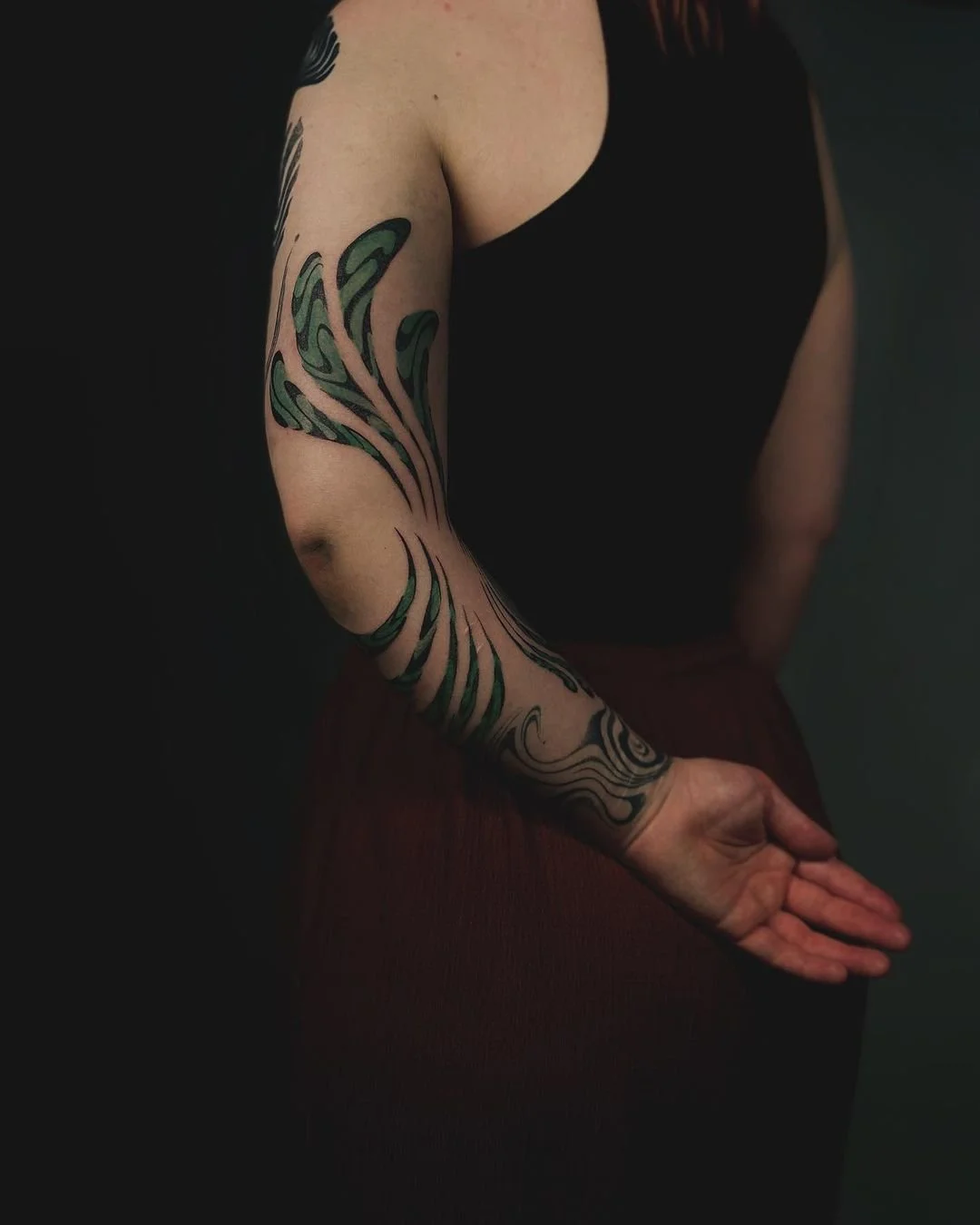 Close-up of a woman's arm with intricate green and black tattoo design, wearing a sleeveless top and a brown skirt, against a dark background.