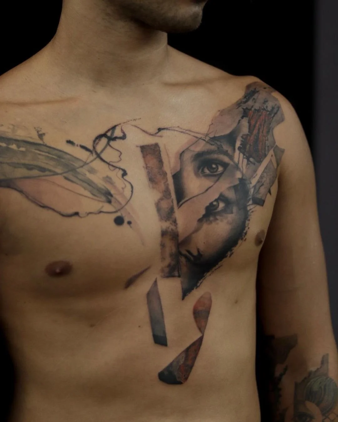 A man's torso featuring a tattoo with an abstract design, including fragments of a face and artistic elements, on the chest.