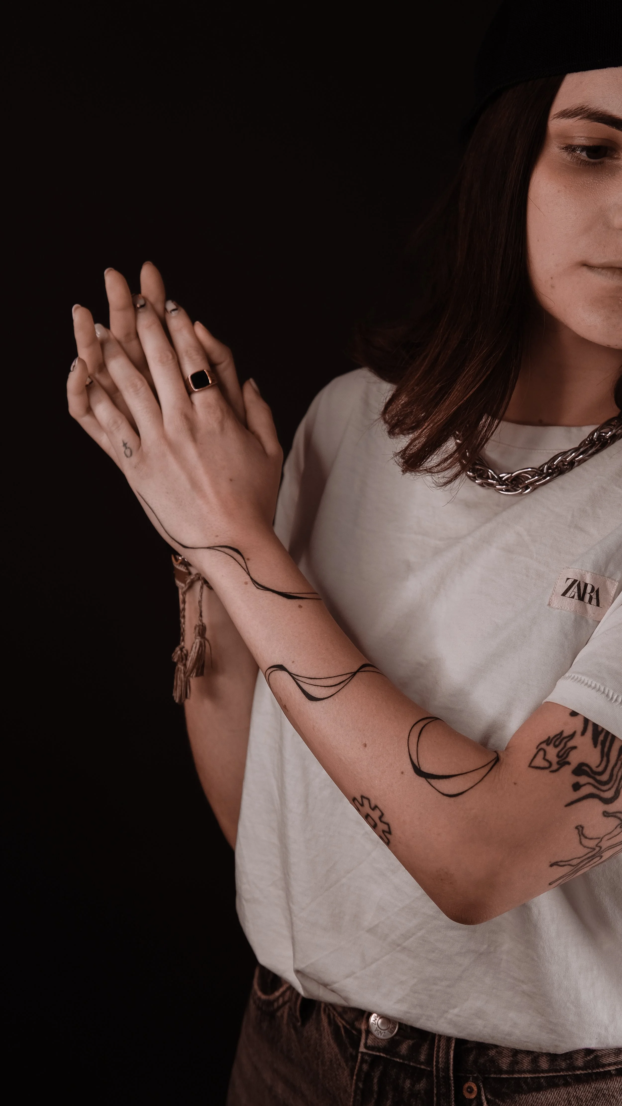 Person wearing a white shirt with tattoos on their arms and hands, including a gear and a crescent moon. They have a ring, a chain necklace, and are clasping their hands.