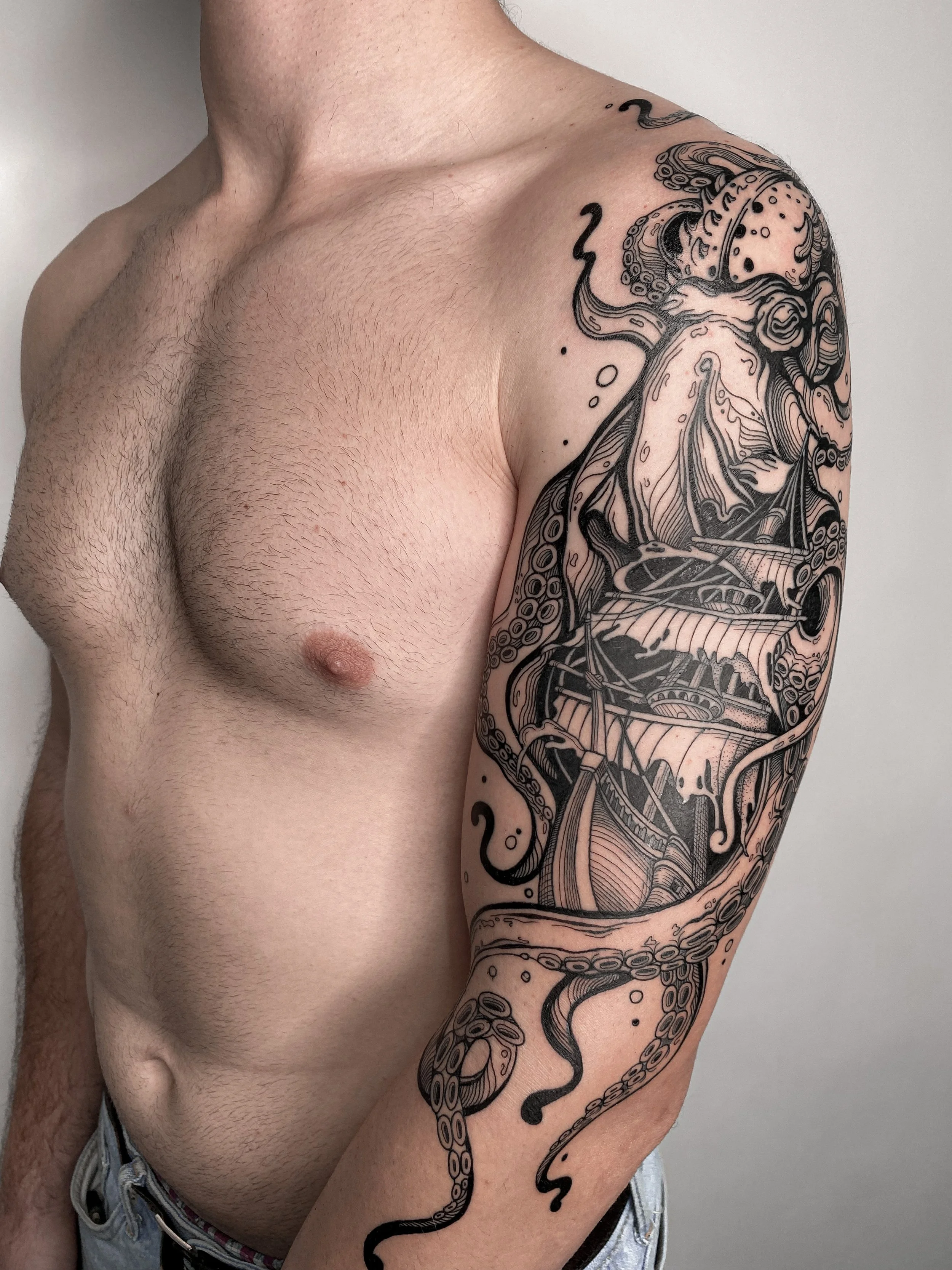Close-up of a tattooed man with a sleeve tattoo on his right arm featuring a detailed, black and grey design of an octopus with a ship in its tentacles, on a plain white background.