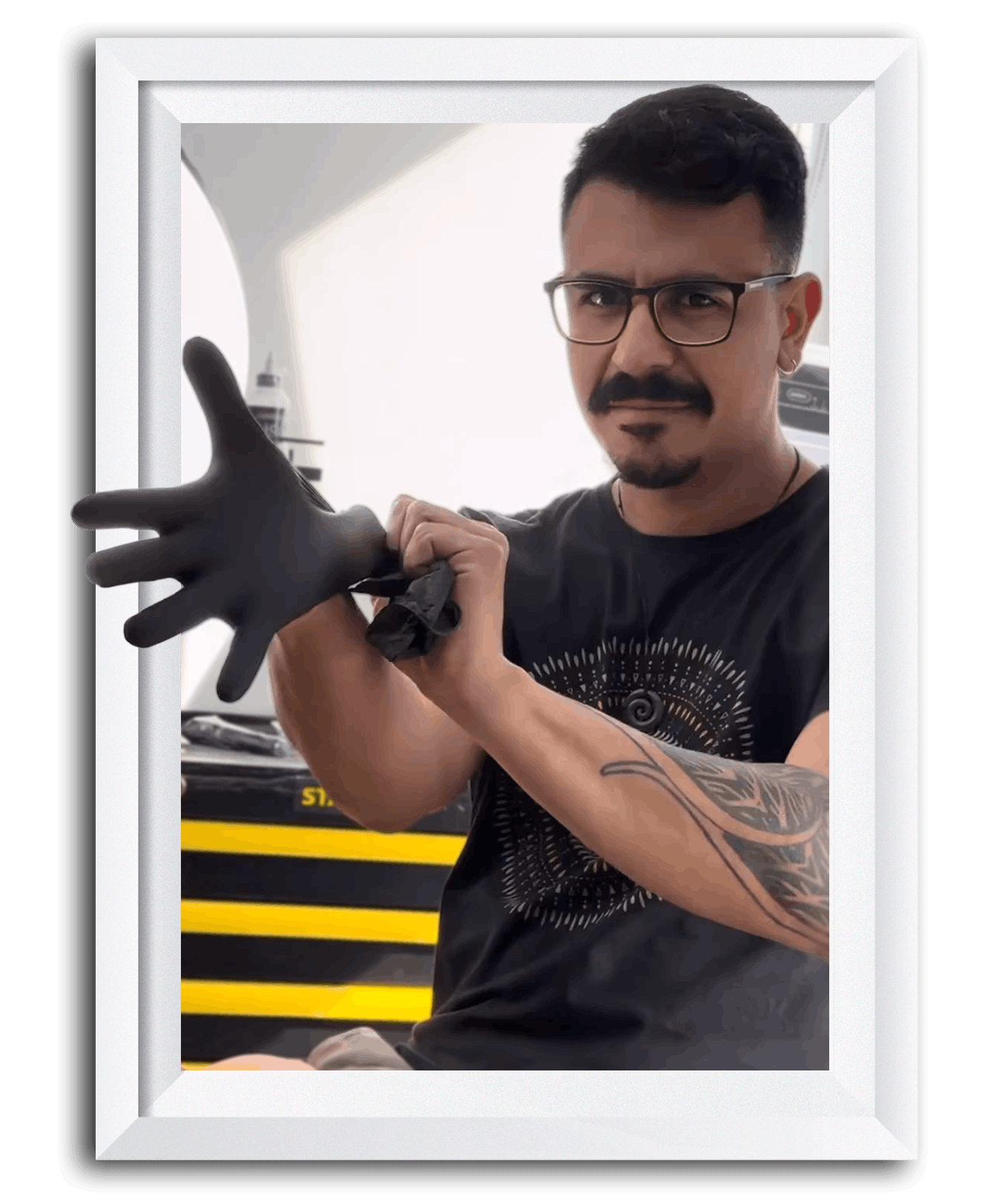 A man with glasses, a black T-shirt, and a tattoo on his arm, putting on a black glove.