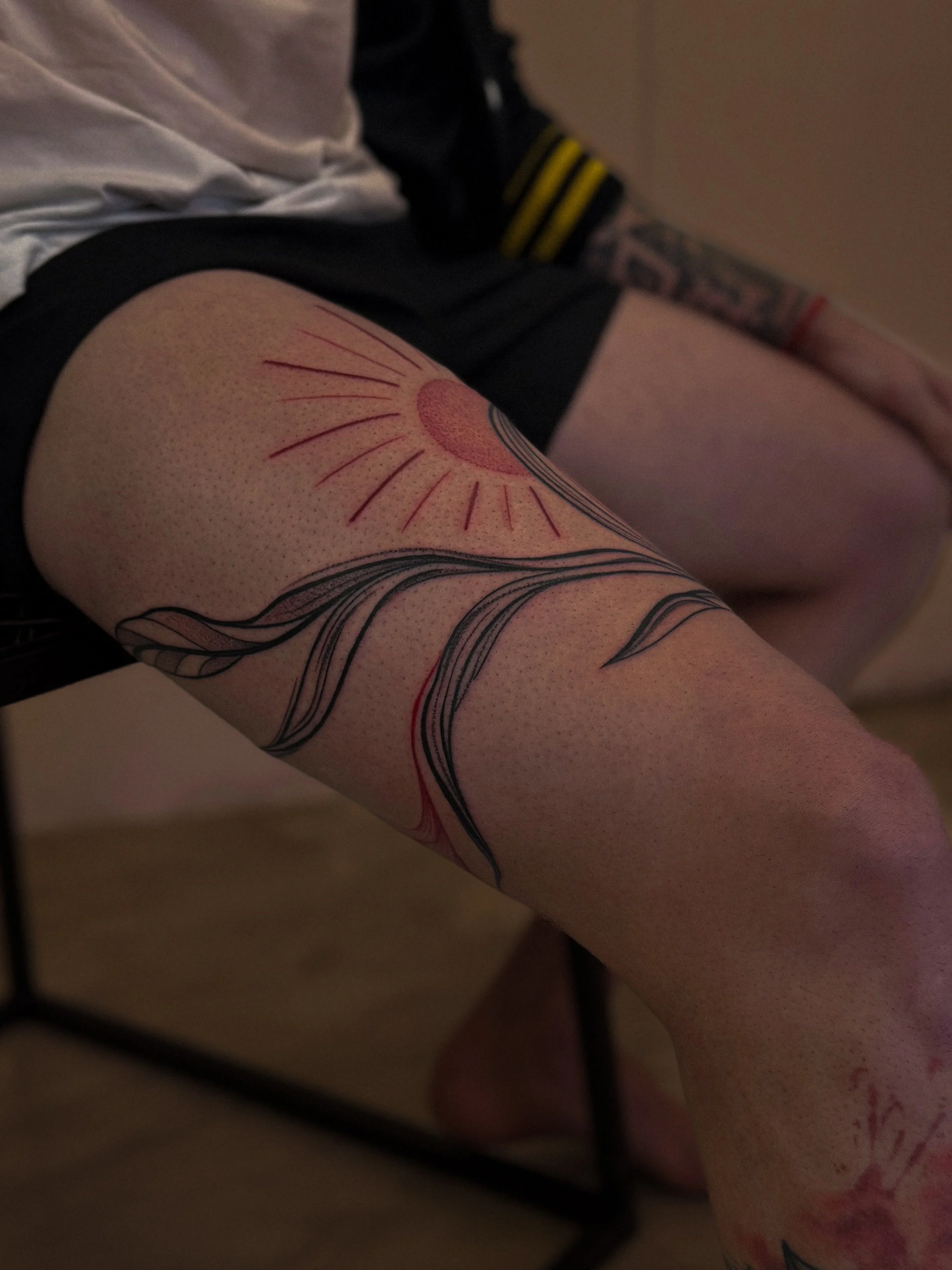 A person's thigh with a tattoo of a sun and flowing black and red lines.