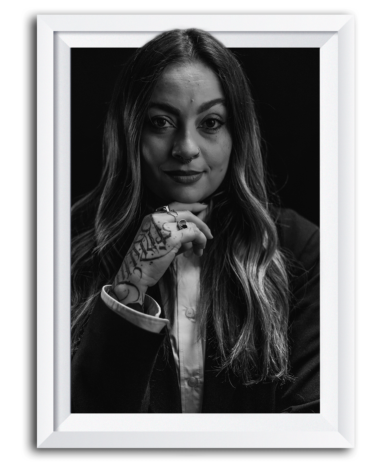Black and white portrait of a woman with long wavy hair, wearing a dark blazer and a white shirt, looking directly at the camera, with tattoos on her hand and neck, framed as if in a picture on a wall.