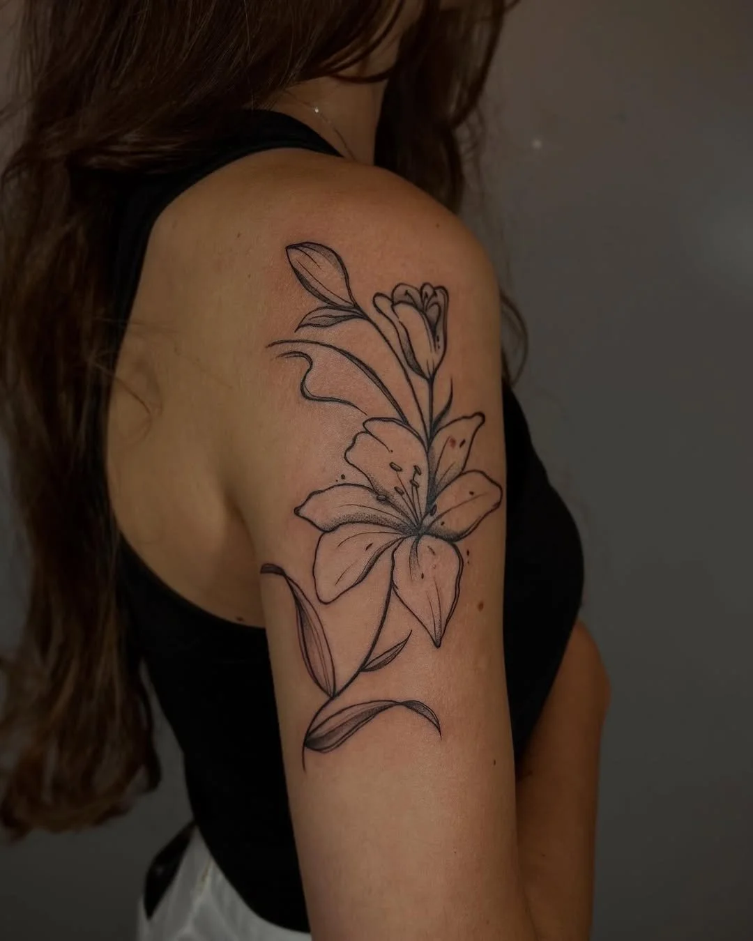 Black line-drawn floral tattoo on woman's shoulder and upper arm, featuring a large lily flower with leaves.