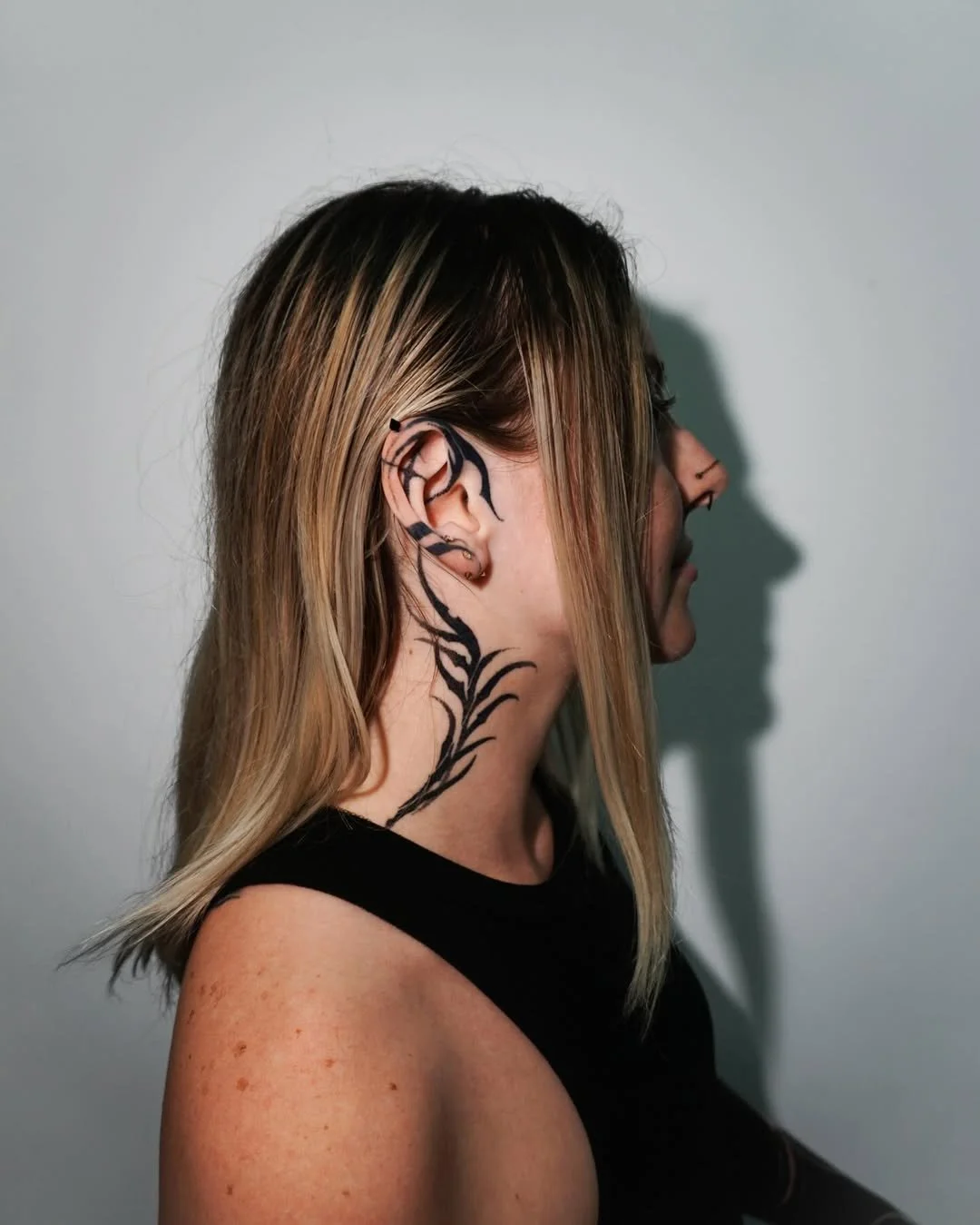 A woman with blonde hair and a black tattoo on her neck and ear, facing sideways against a plain background.