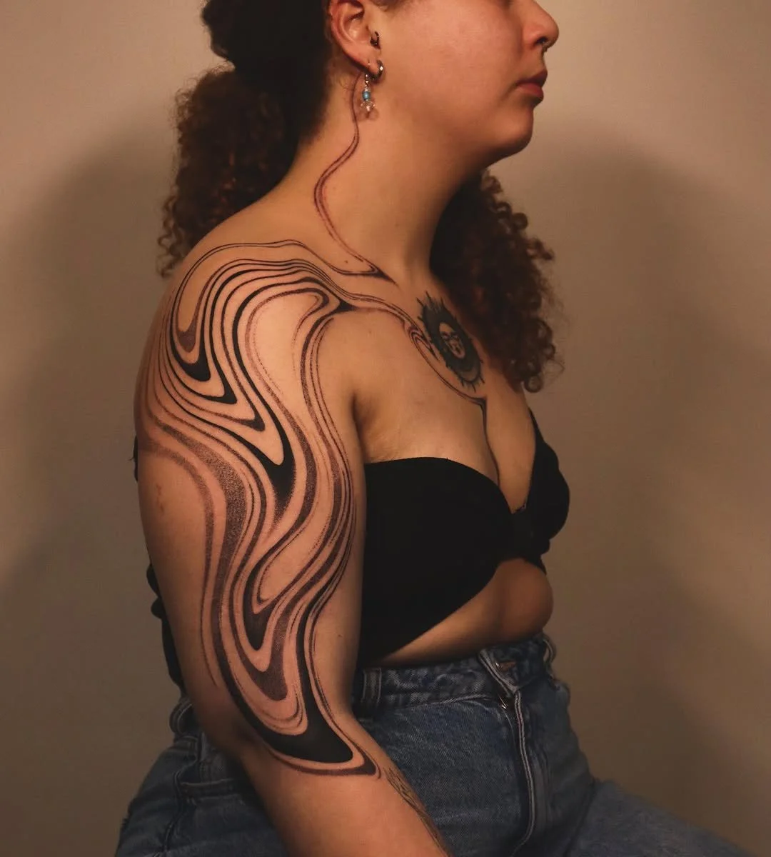 Person with intricate arm tattoo in black and beige, wearing a black top and blue jeans, with curly hair and earrings.