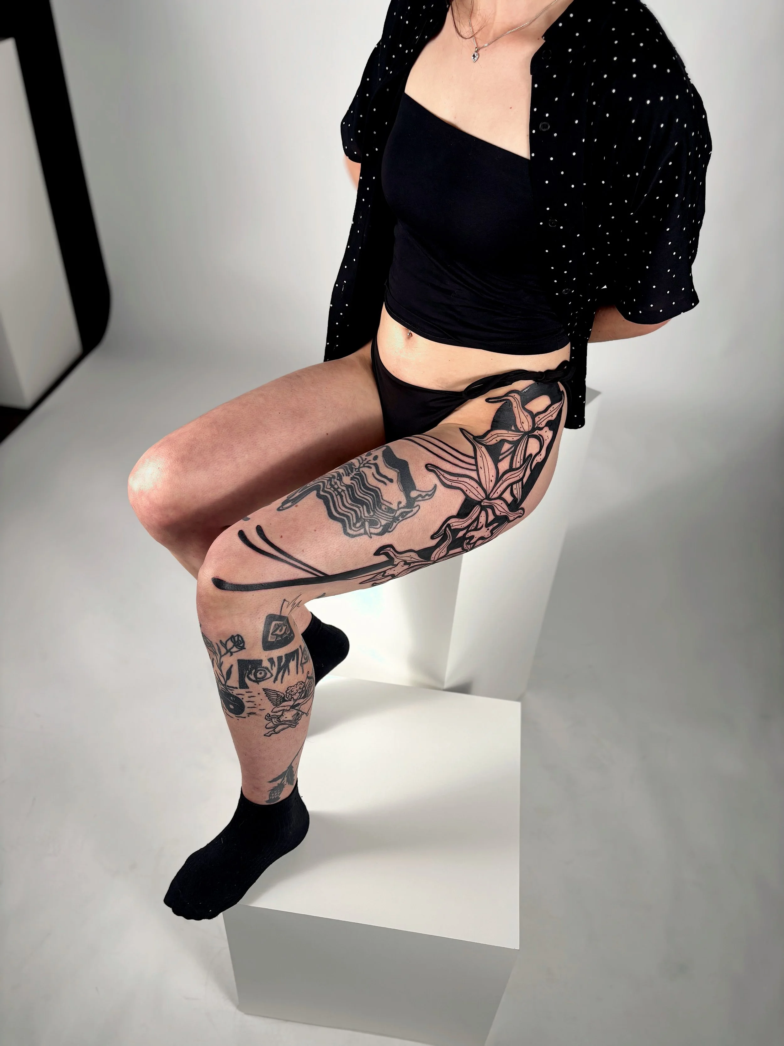 A woman sitting on a white block with tattoos on her right leg, wearing black shorts, a black top, black socks, and a black polka-dot shirt, against a plain gray background.