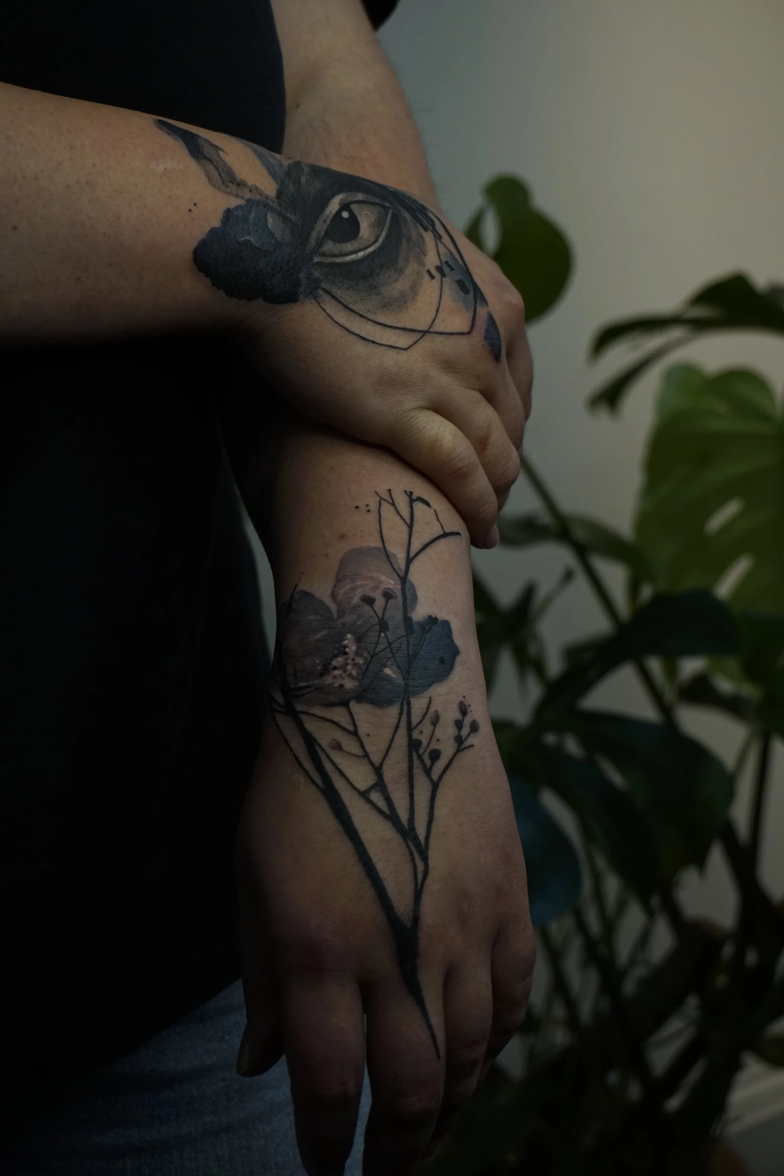A person's arm with several tattoos, including a detailed eye and bird on the upper arm and a floral illustration with watercolor accents on the forearm, with indoor plants in the background.