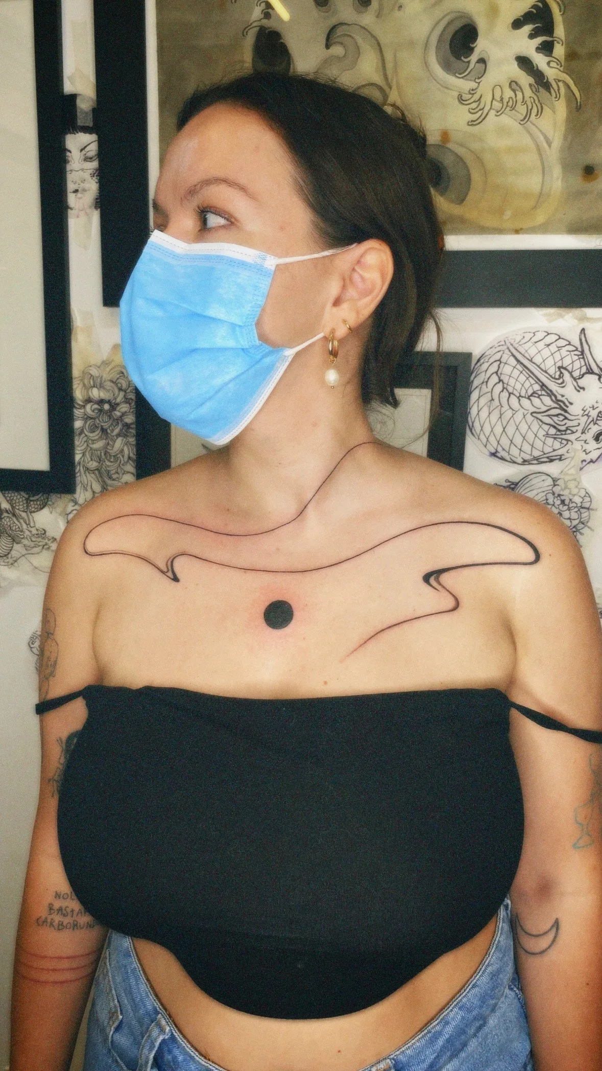 A woman with dark hair and multiple tattoos, wearing a blue surgical mask, a black off-shoulder top, and blue jeans, stands in front of tattoo artwork.