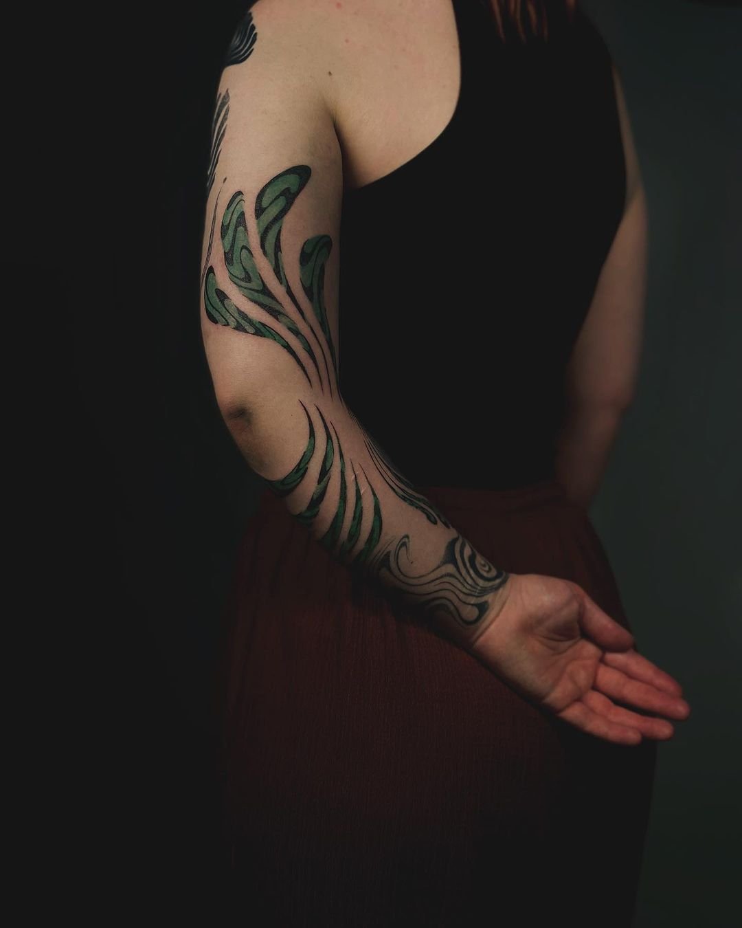 Person with green and black abstract tattoo on left arm, wearing black top and brown skirt, against dark background.