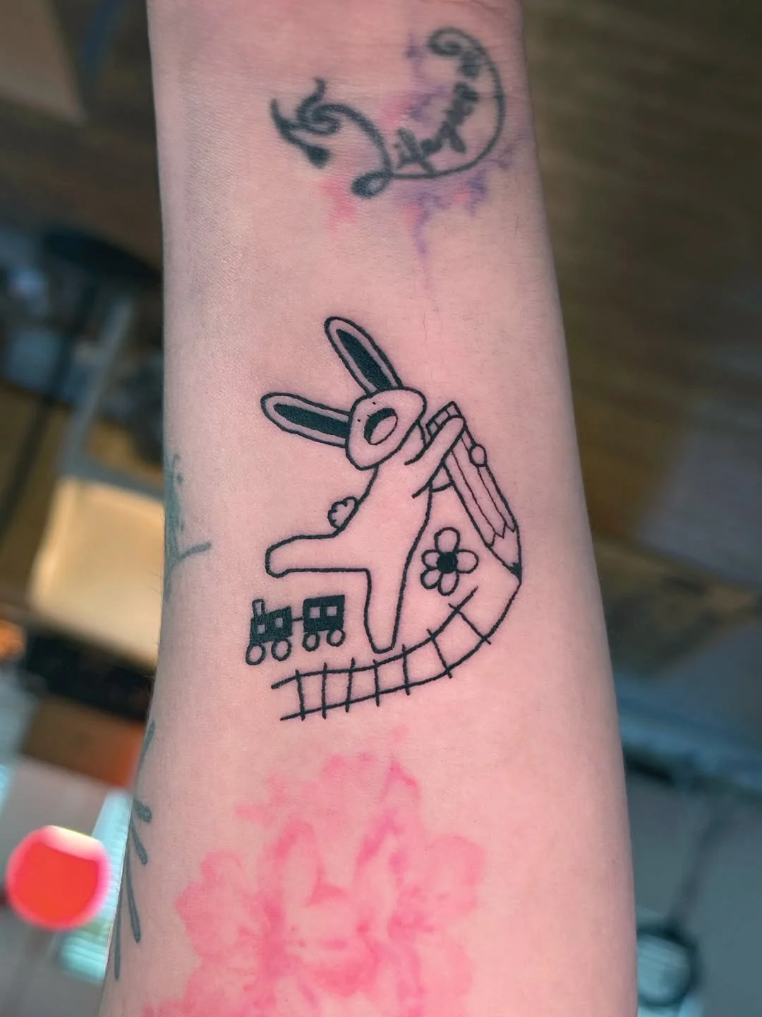 A tattoo of a bunny with a flower on its chest, holding a paintbrush, with a train underneath and a curved line resembling a rainbow or a roller coaster beneath the train.