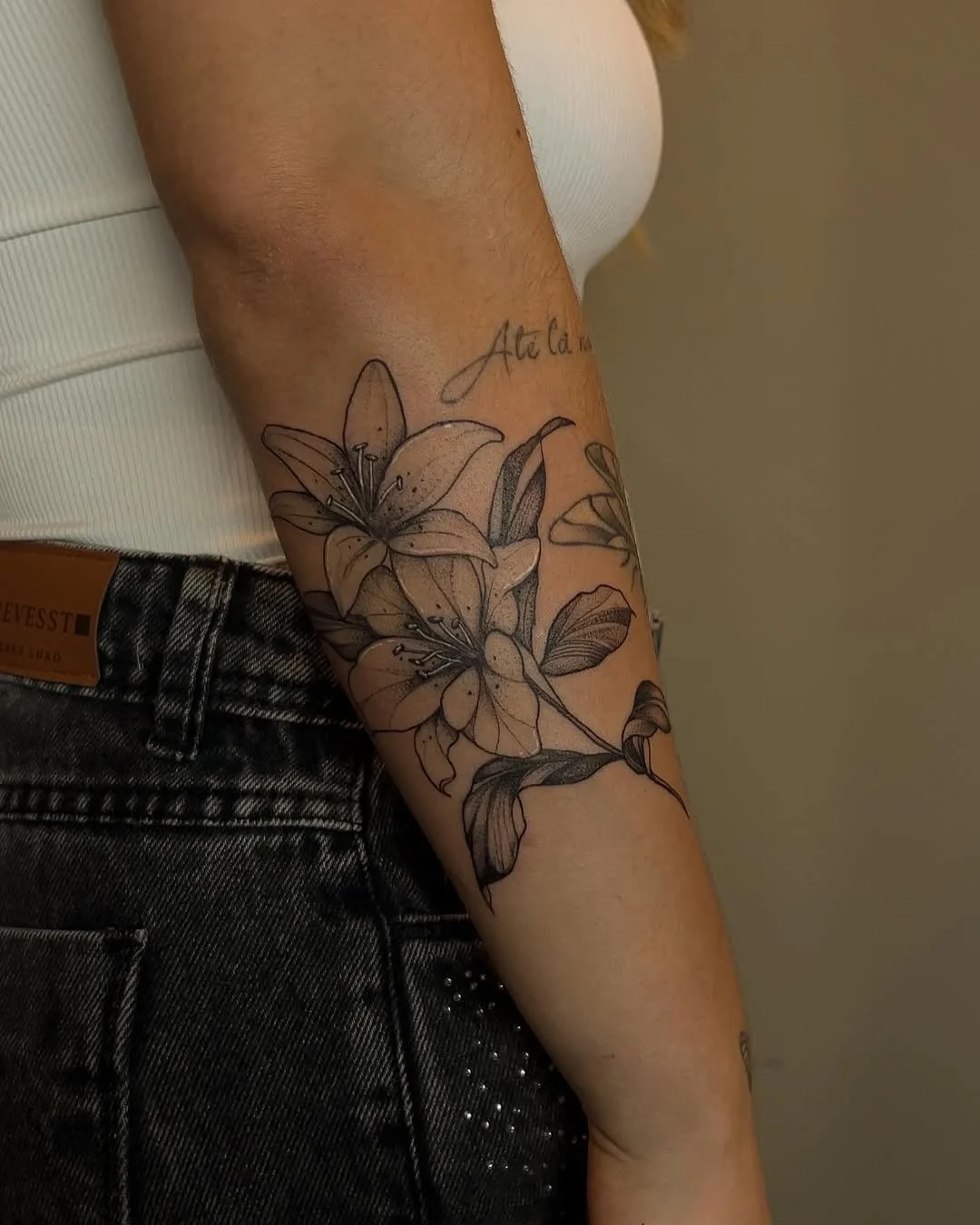 Black and gray tattoo of lilies with leaves on a person's forearm, with cursive script above.