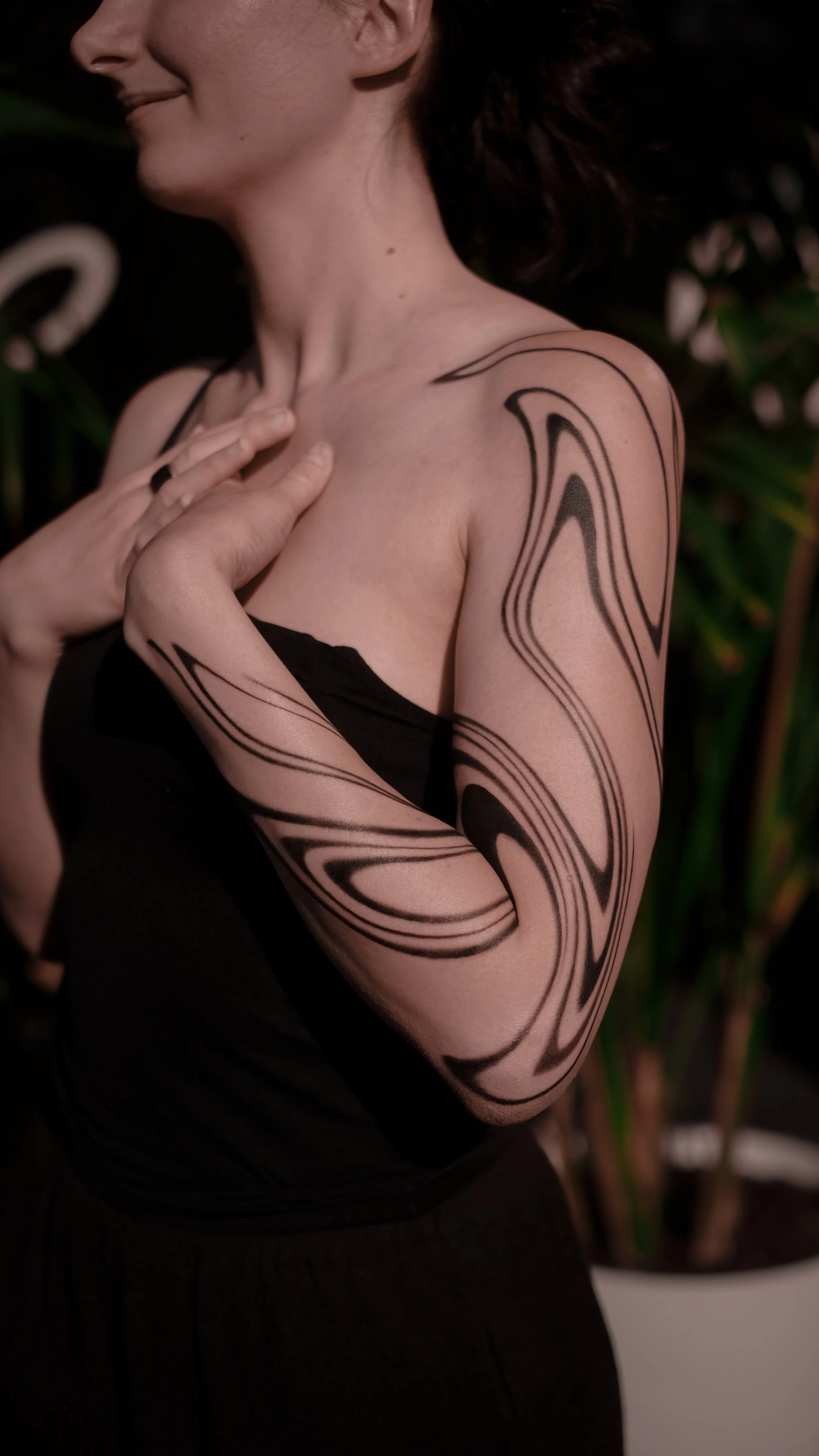 Woman with abstract arm tattoo, wearing black, standing against a plant background.