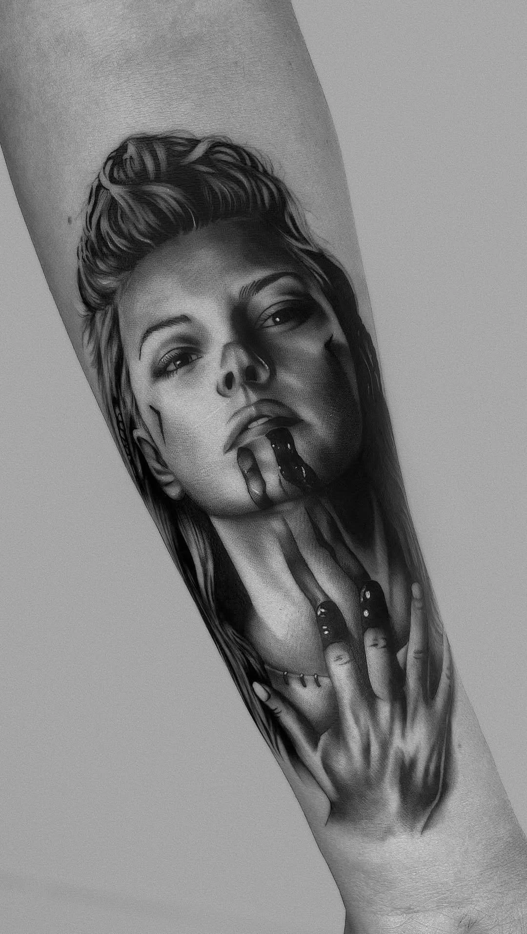 Black and gray tattoo on forearm depicts a realistic portrait of a woman with long hair, looking upward and touching her face with her hand.