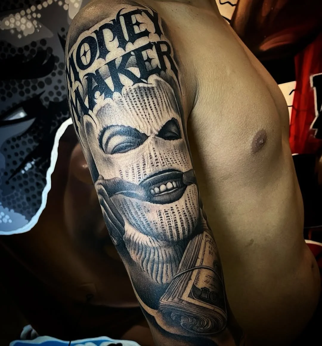 Tattoo of a smiling face with the words 'MONEY MAKER' at the top, holding a bundle of cash, on a person's upper arm.