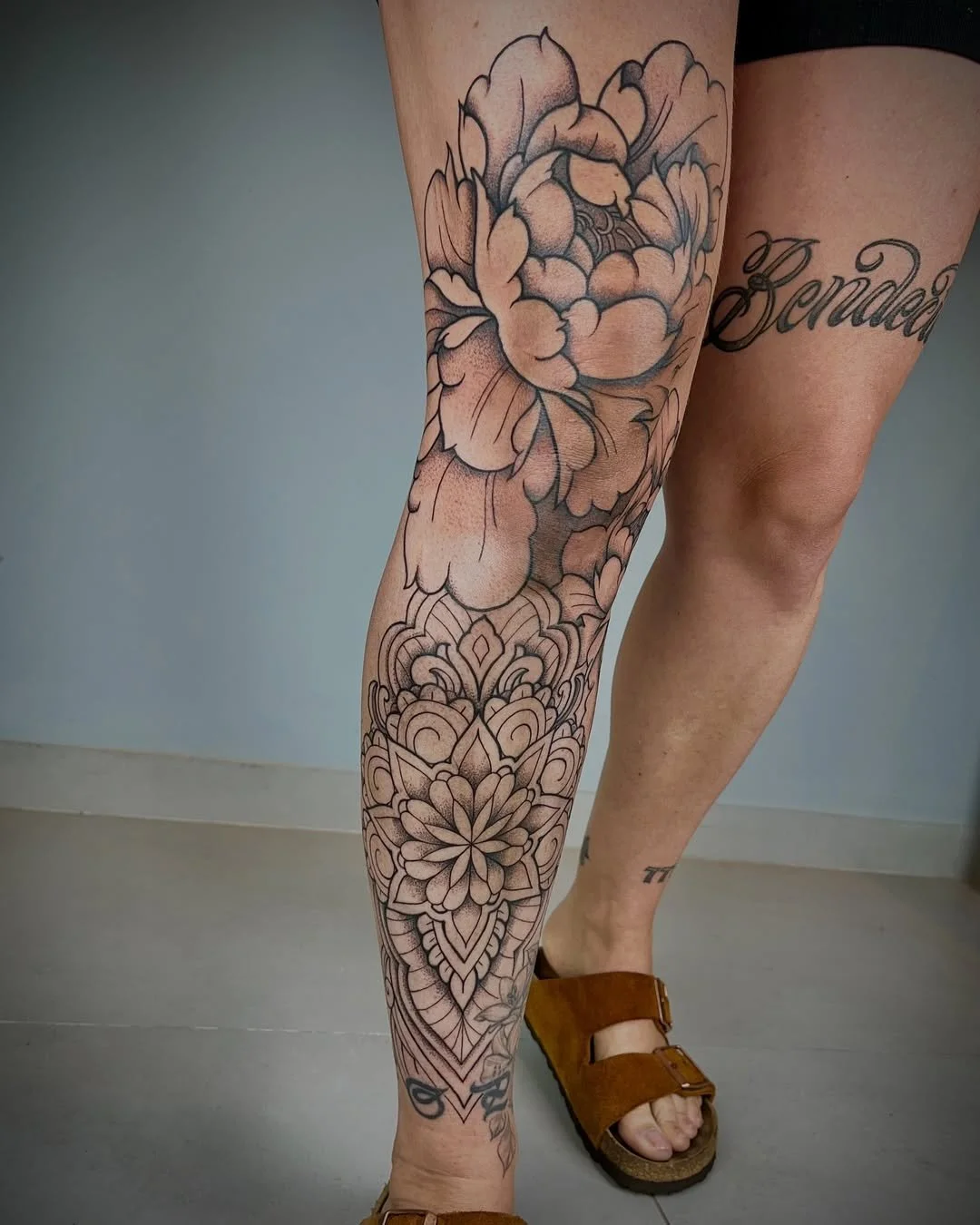 Tattoo of a large flower with intricate patterns on the lower leg, and part of the word 'Canada' in cursive on the upper leg.